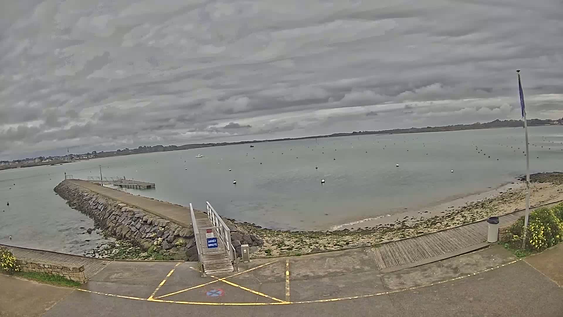 A calm body of water under a cloudy sky is seen from a parking lot next to a stone jetty and wooden ramp leading to a small dock.