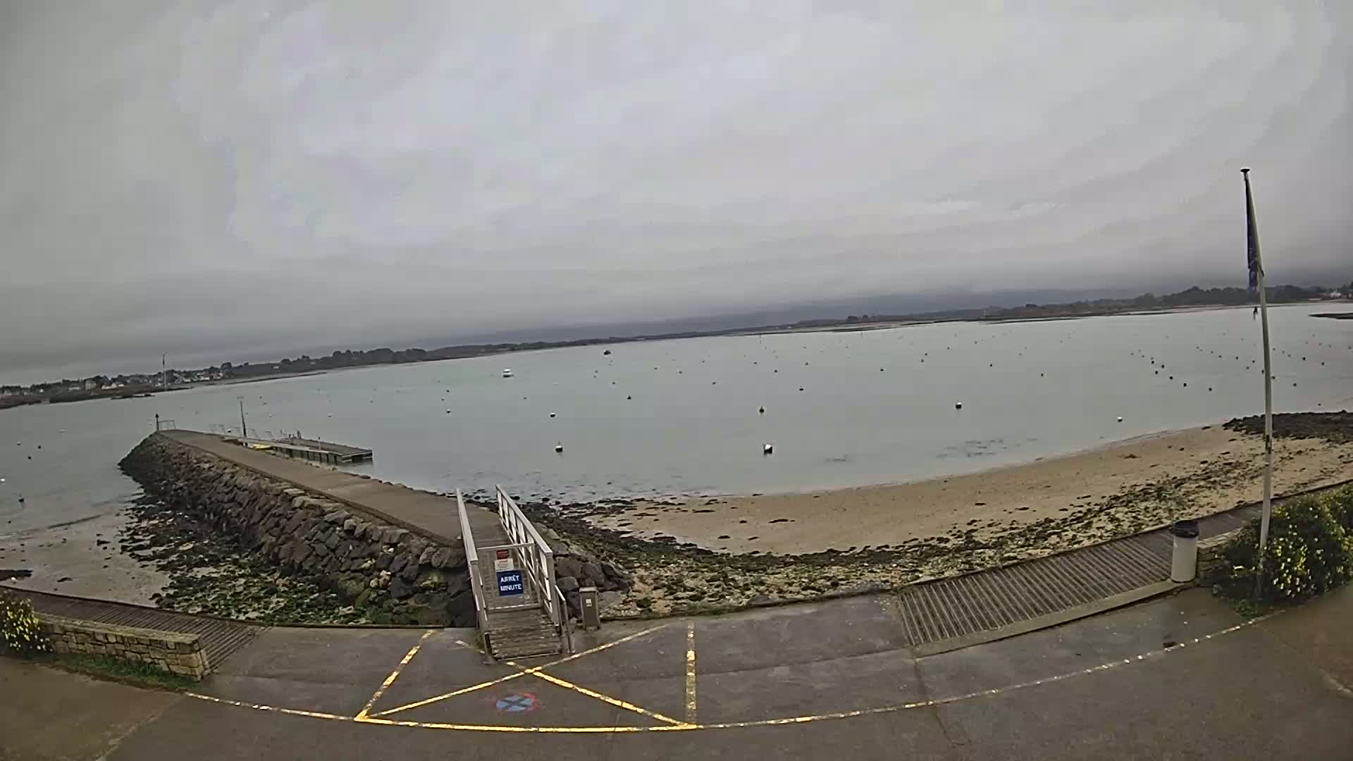 A stone jetty extends into a calm, gray-sky body of water dotted with buoys, leading to a small sandy beach.