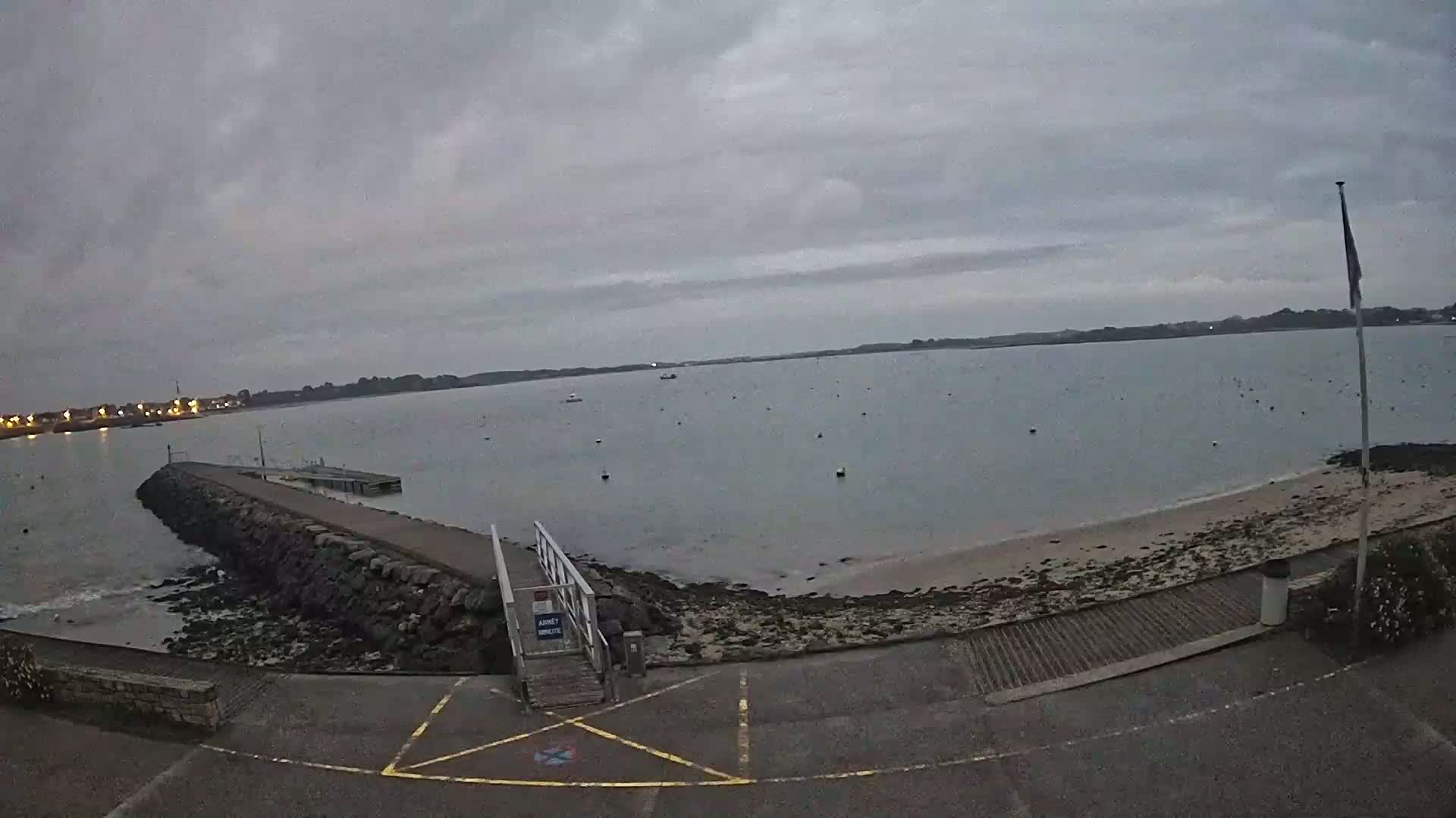 A calm, overcast day reveals a rocky shoreline with a small pier and boat ramp, overlooking a bay dotted with buoys.
