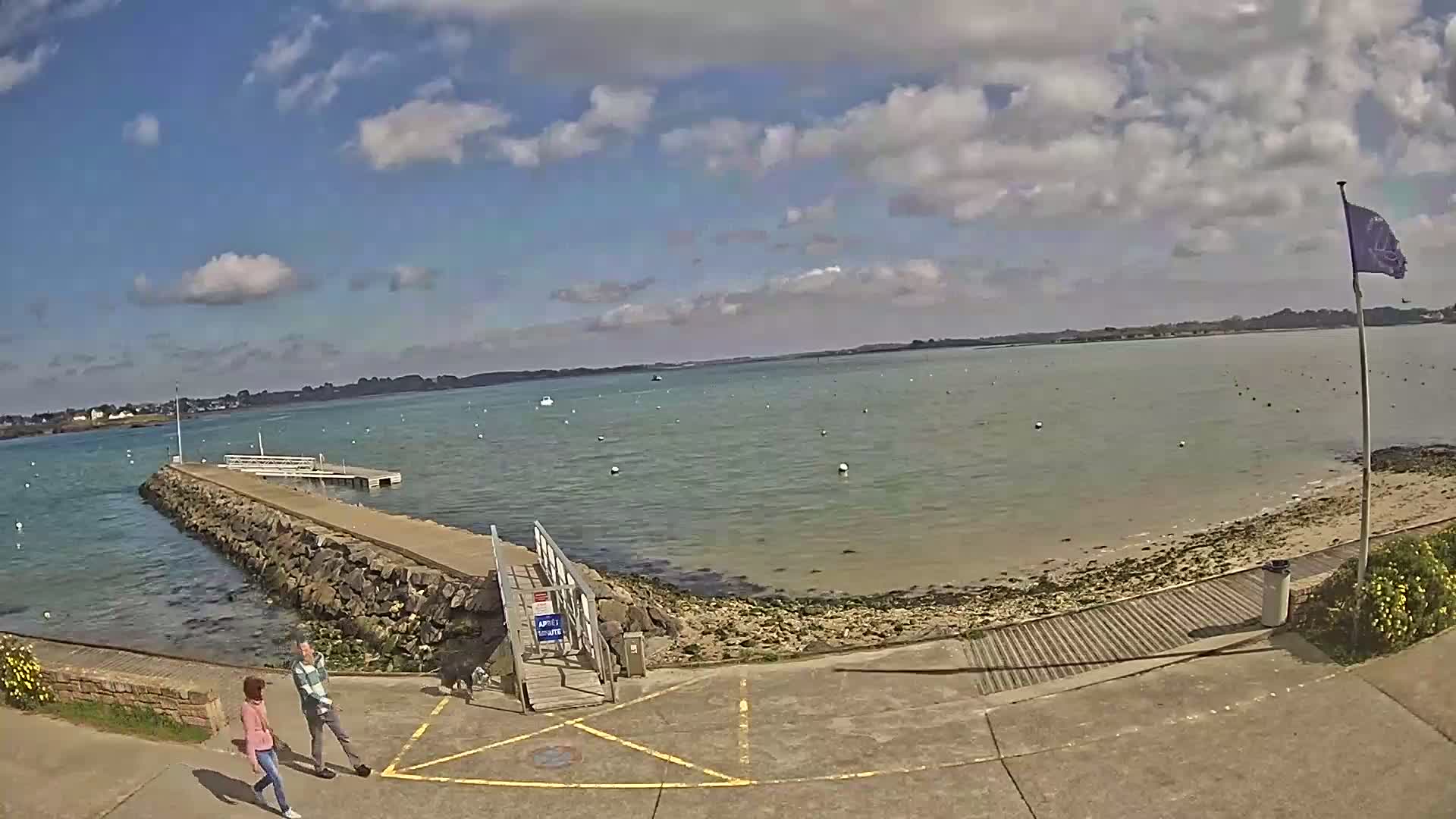 A partly sunny day at a waterfront area with a small dock, a sandy beach, and two people walking near a metal railing.