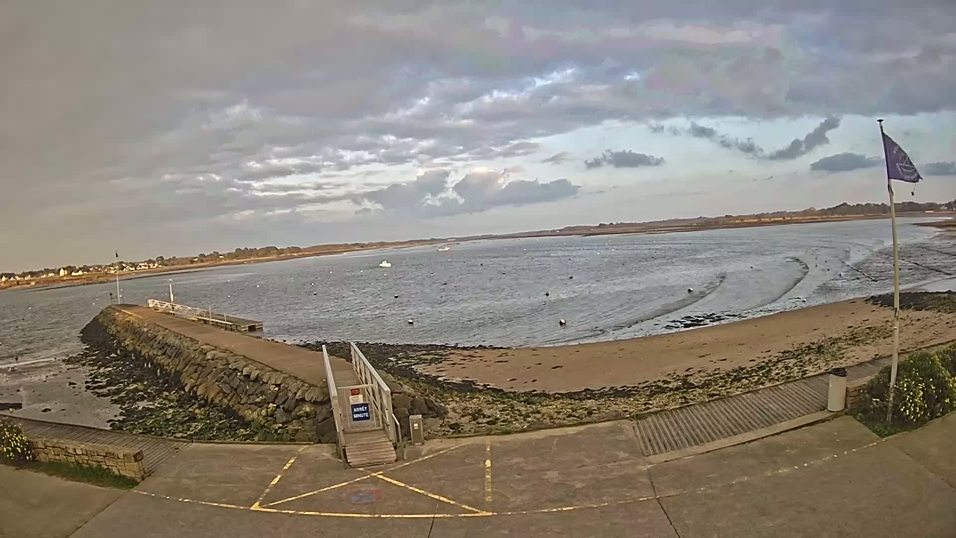A stone jetty extends into a calm body of water under a cloudy sky, with a sandy beach and a small town visible in the distance.