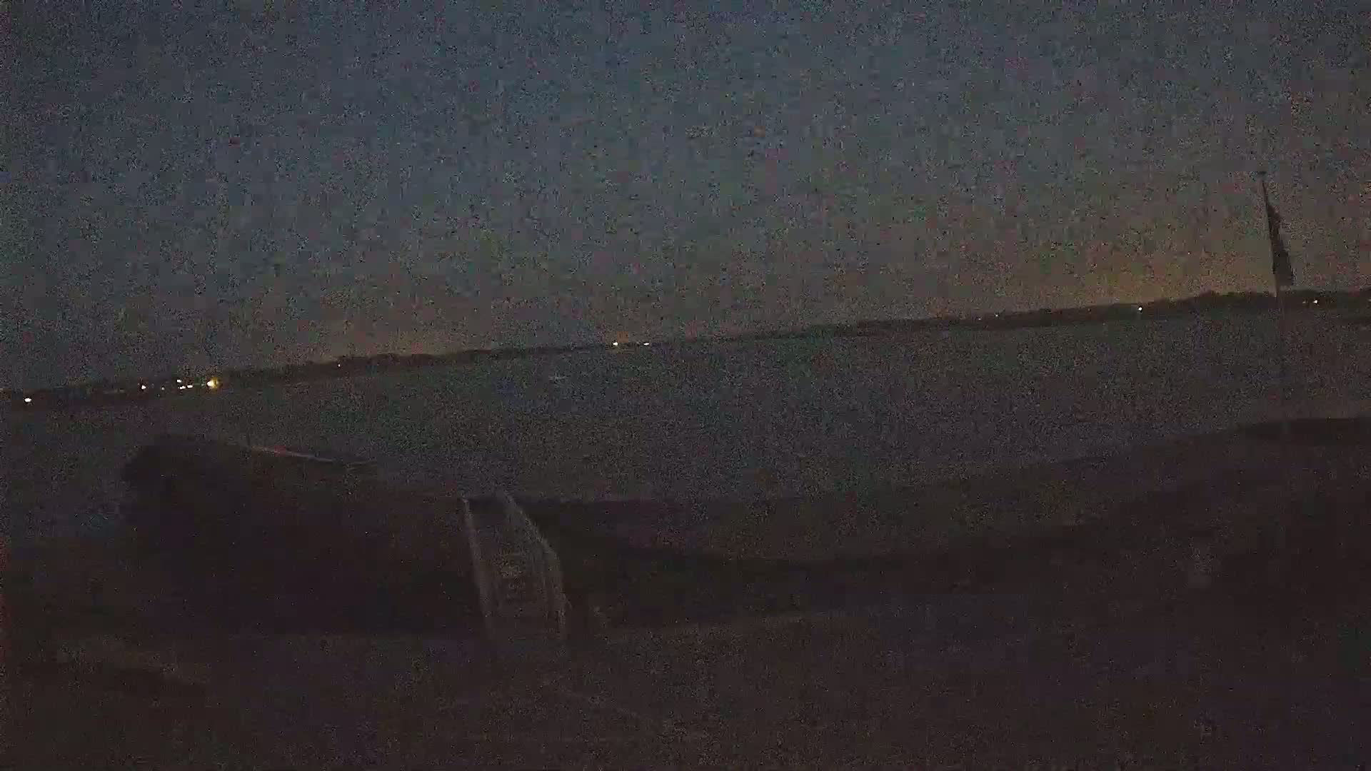 A dark, nighttime scene shows a body of water under a mostly clear, dark sky with a few distant lights visible on the horizon.