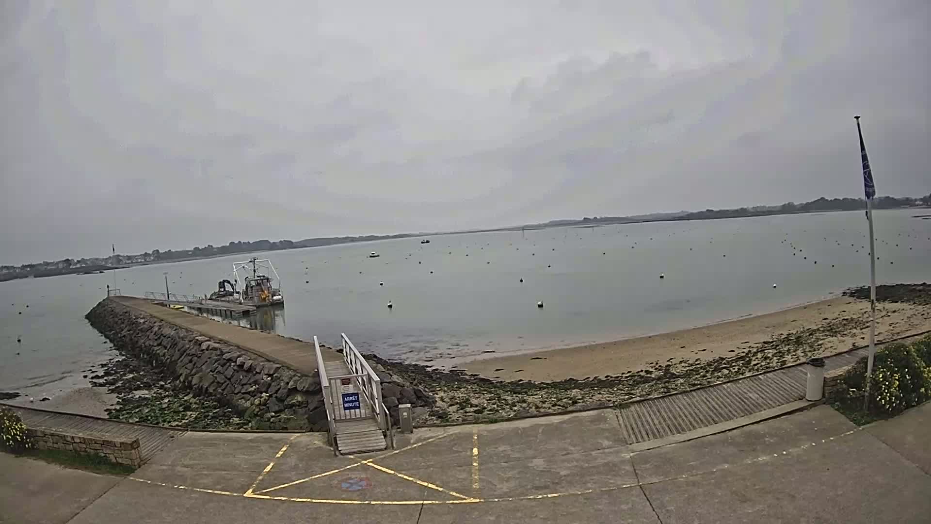 A calm, overcast day reveals a small boat docked at a stone jetty leading to a sandy beach with numerous buoys in the water.