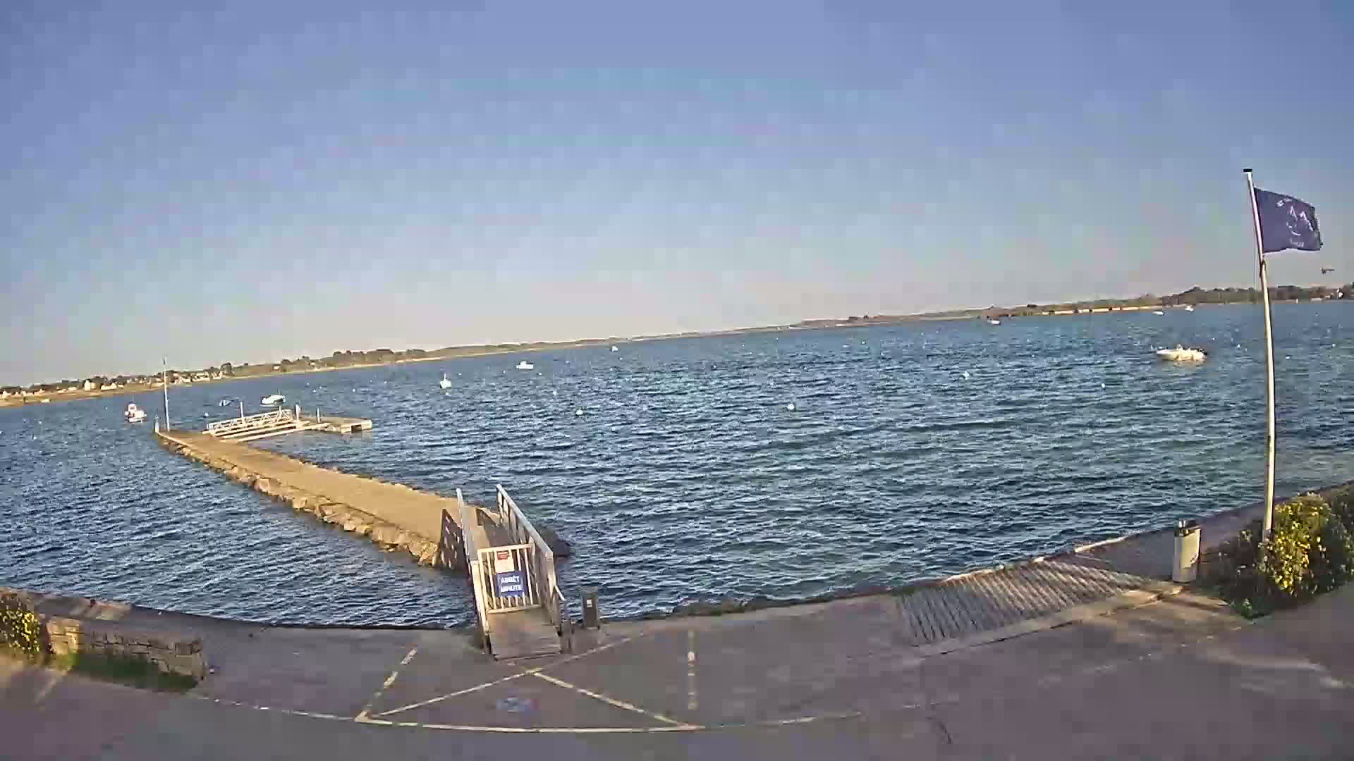 A calm, sunny day reveals a small boat launch extending into a body of water with several boats scattered across its surface, and a flagpole with a flag stands to the right.