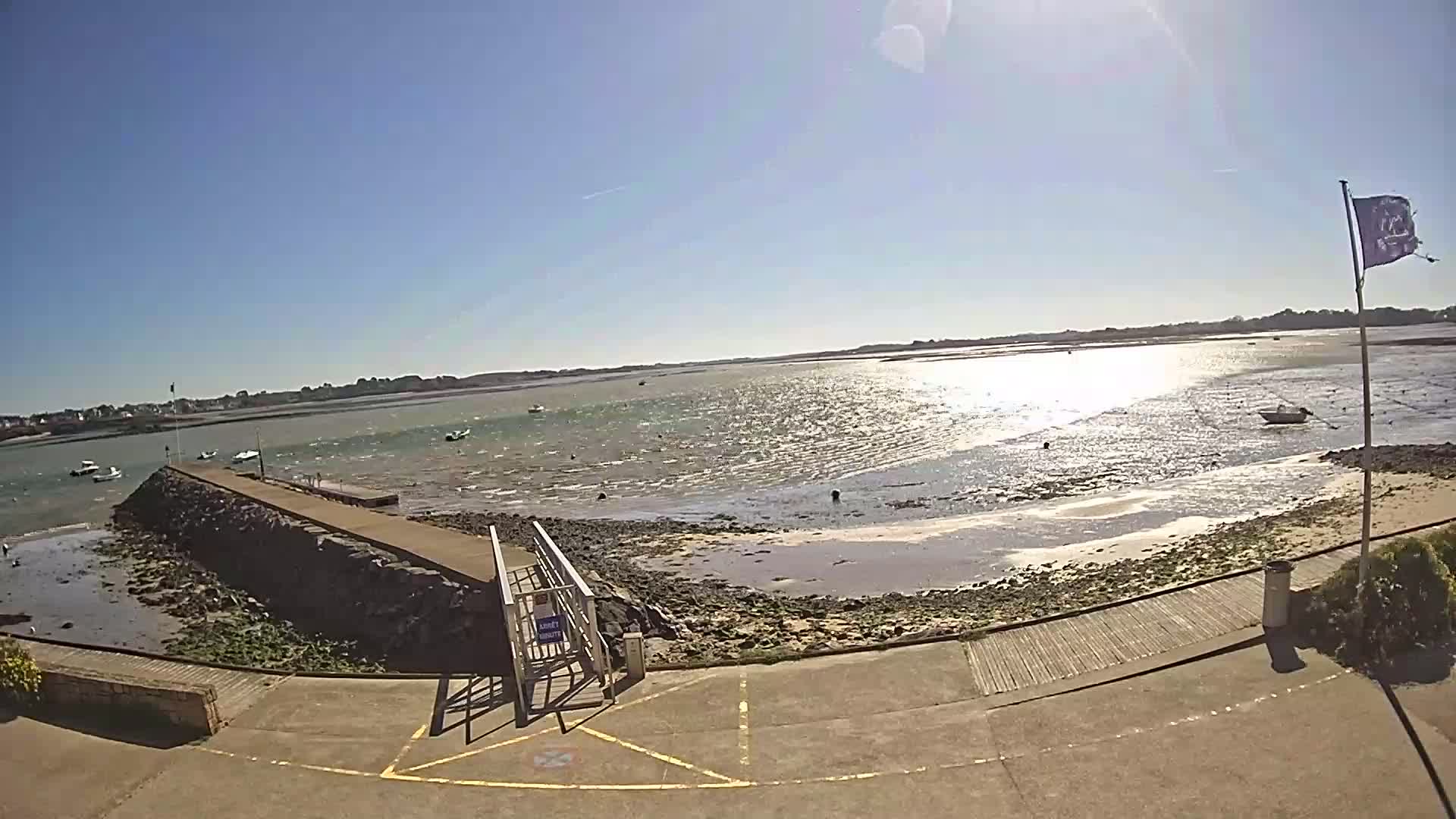 A sunny day shows a calm body of water with several small boats, a stone jetty with a walkway leading to a paved area, and a flagpole with a purple flag.