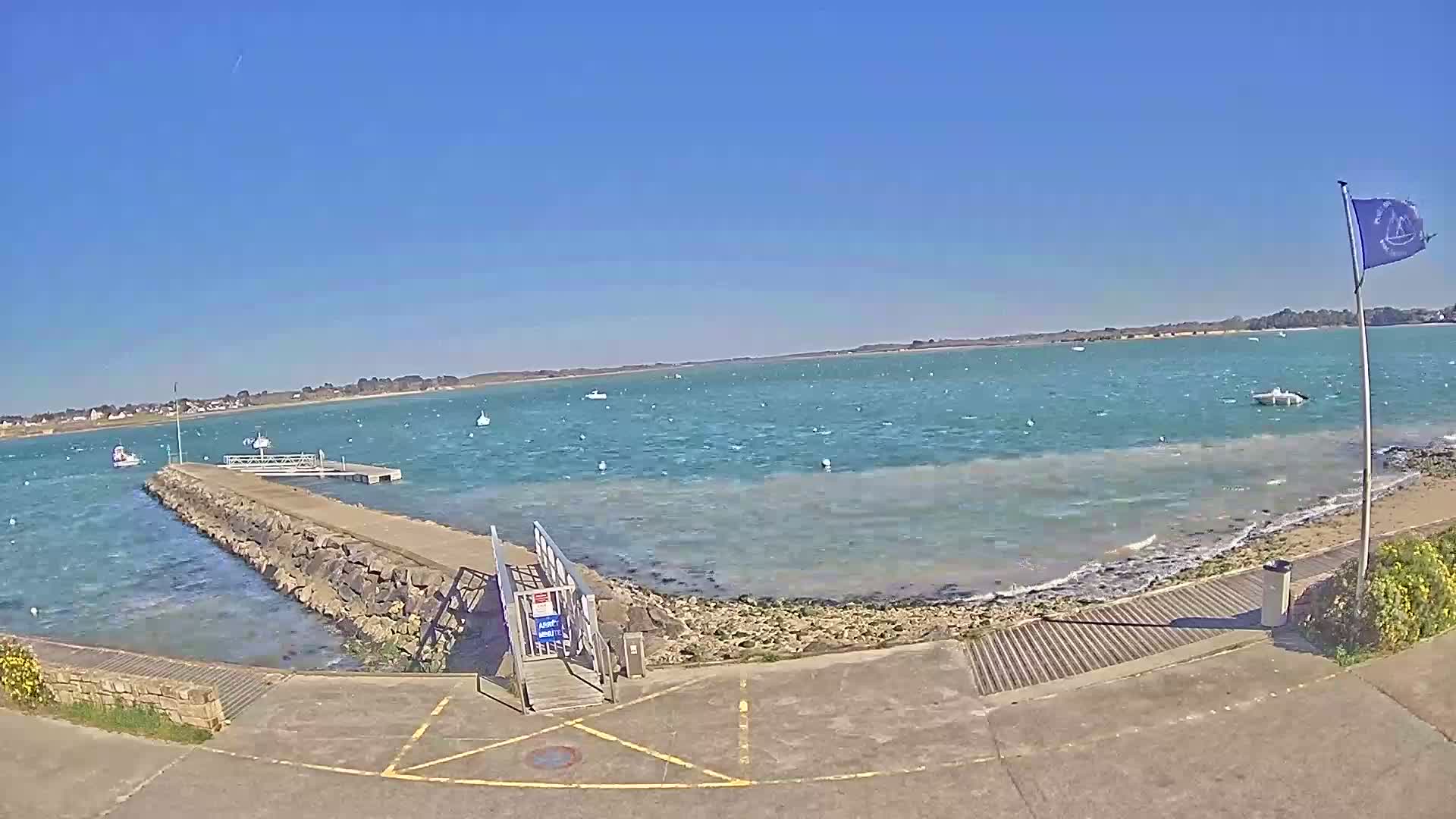 A stone pier extends into a windy, turquoise body of water on a sunny day, with a few small boats visible in the distance.