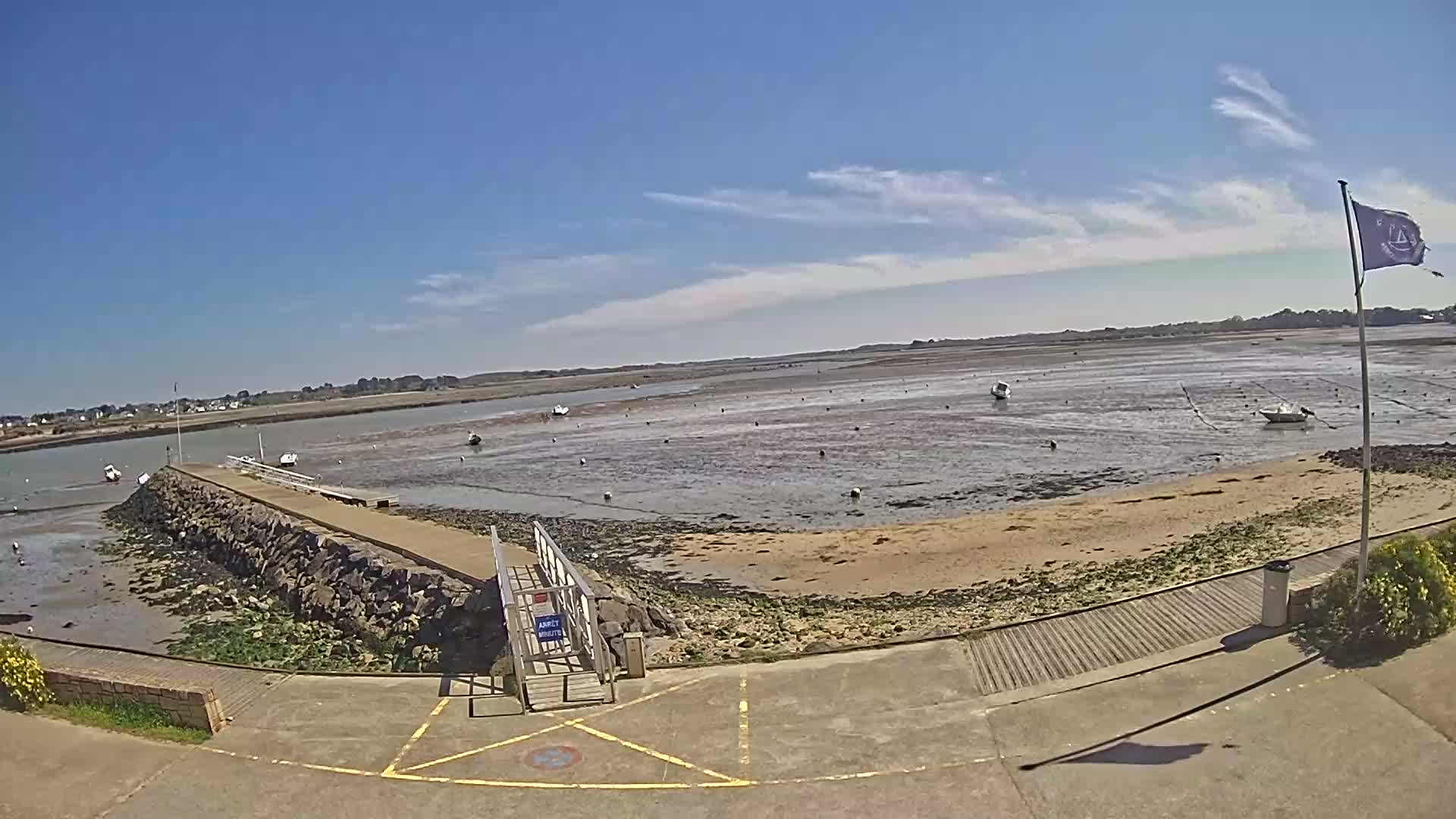 A stone pier extends into a tidal bay under a mostly sunny sky, with several small boats visible at low tide.