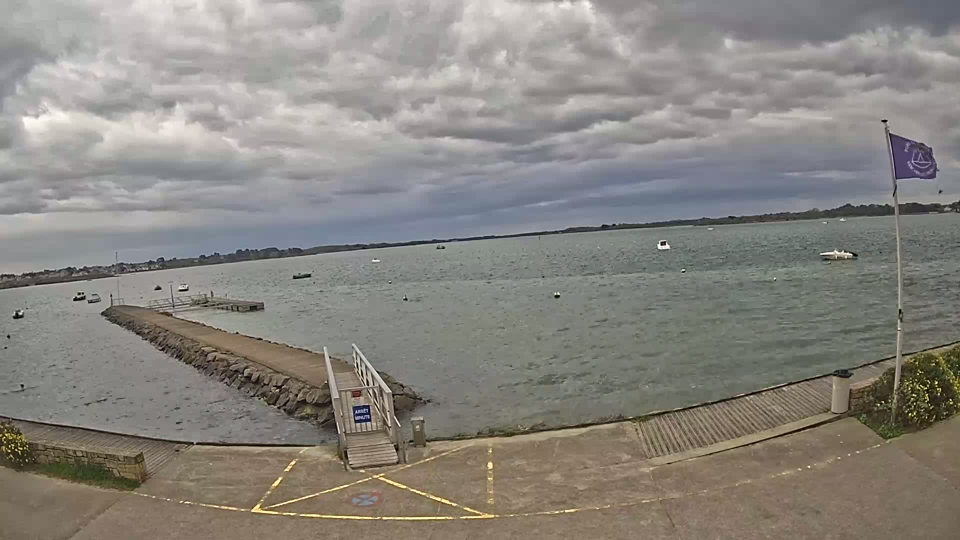 A stone pier extends into a calm body of water under a cloudy sky, with several small boats scattered across the bay.