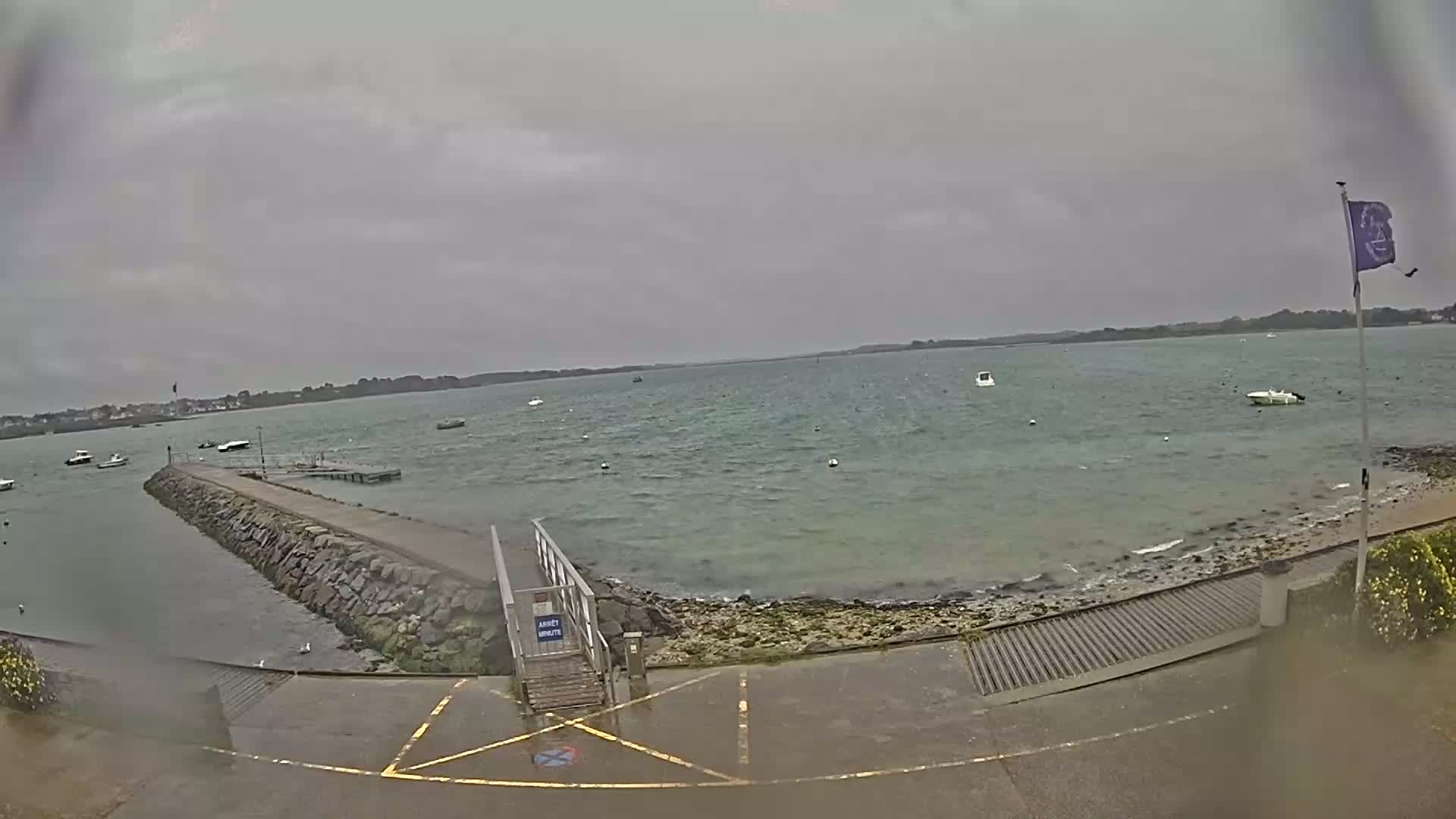 A mostly cloudy day shows a calm body of water with several small boats, a stone jetty leading to a small dock, and a paved area with a ramp.