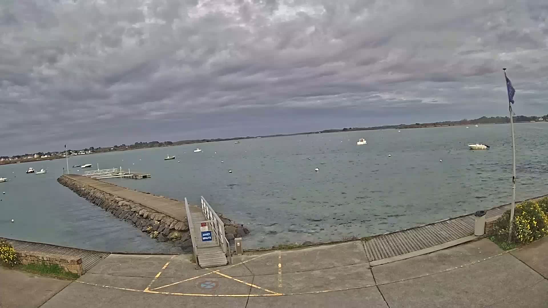 A stone jetty extends into a calm body of water under a cloudy sky, with several small boats scattered across the surface.
