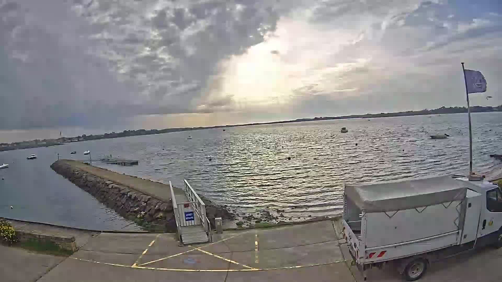 A partly cloudy sky overlooks a calm body of water with a few boats, a small stone jetty, and a parked white truck.