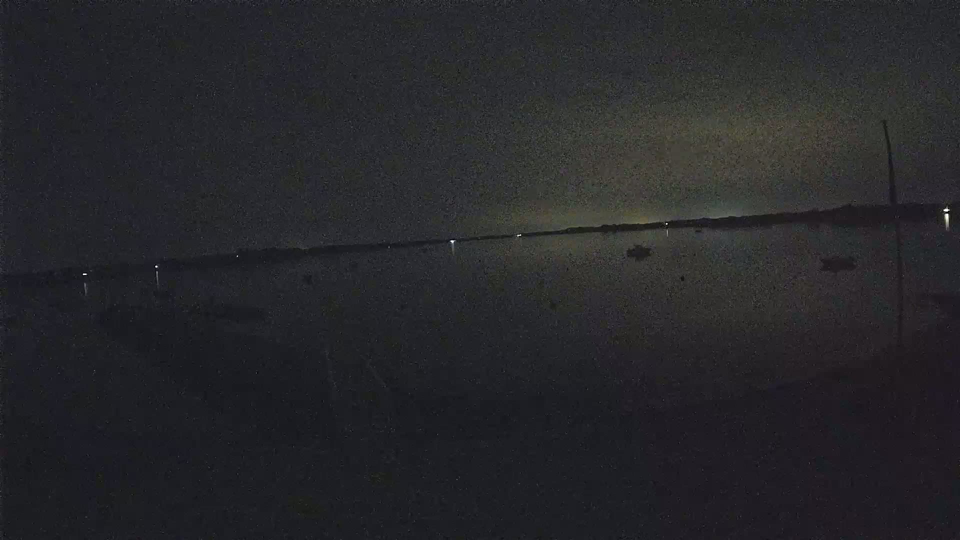A dark, nighttime view of a calm body of water with a few faint lights visible on the far shore and some sparsely scattered lights on the water.