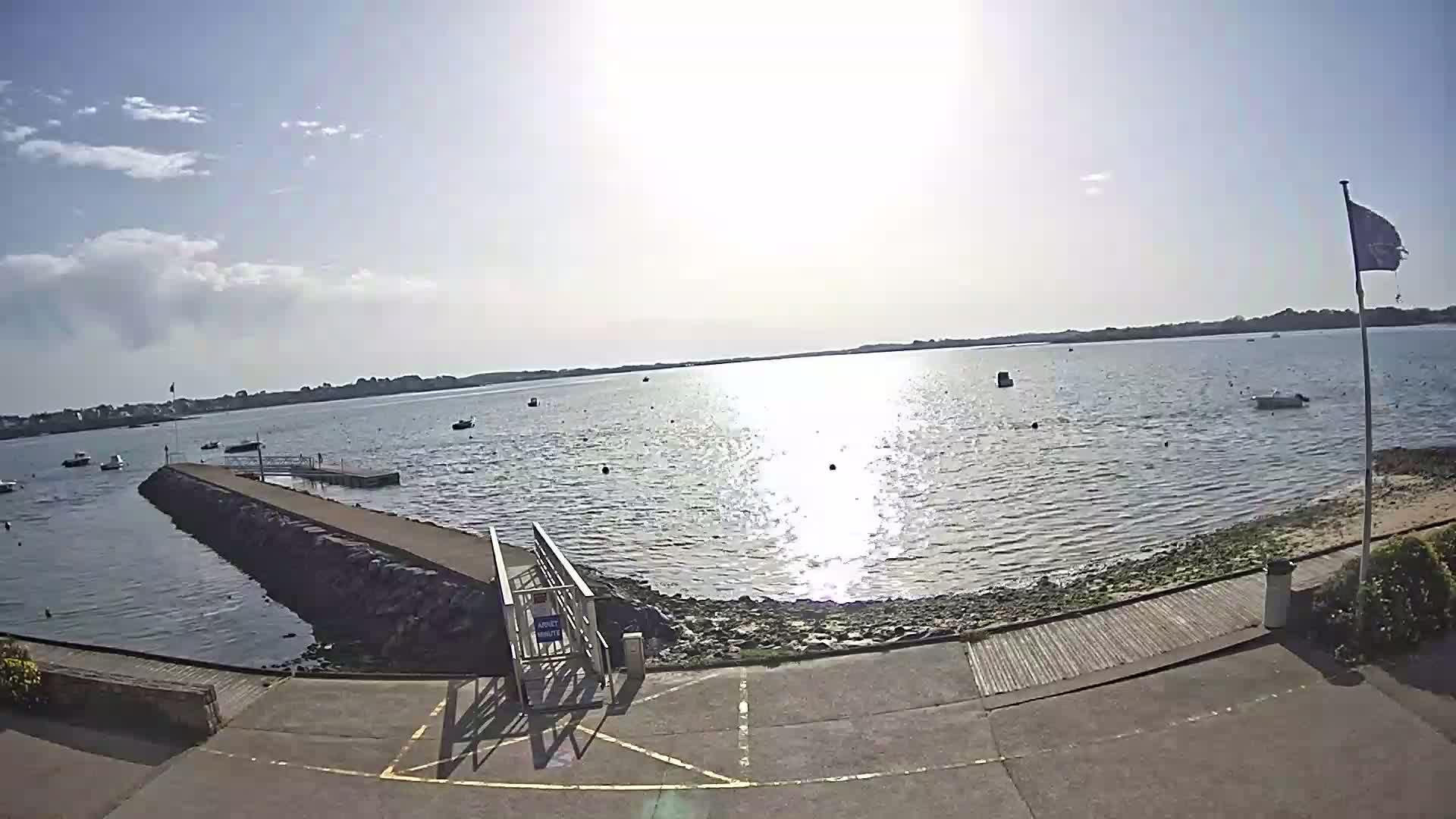 A sunny day reveals a calm body of water with scattered boats, viewed from a paved area leading to a stone jetty.