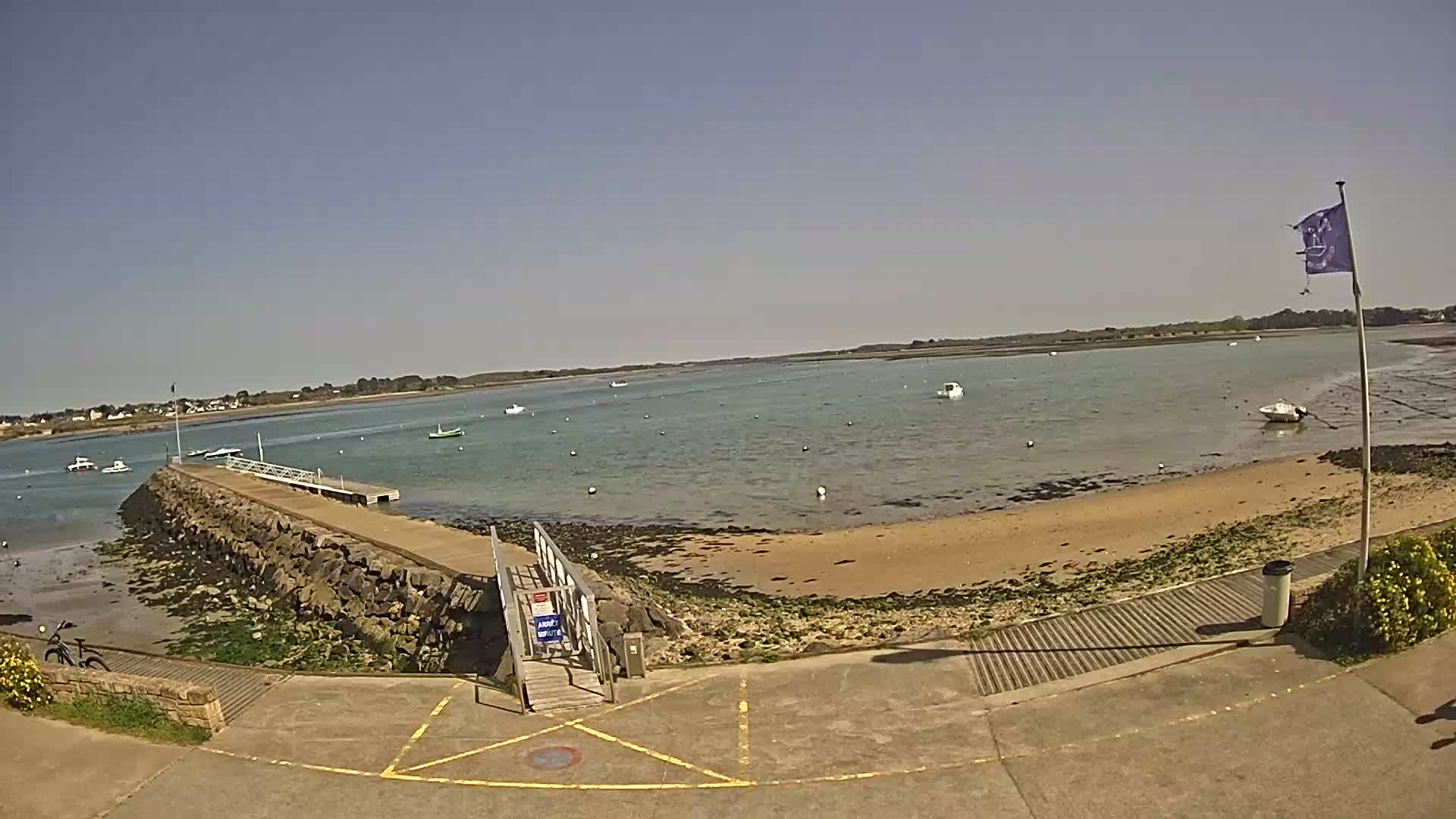 A stone pier extends into a calm, shallow body of water under a clear blue sky, with several small boats visible in the distance and a sandy beach nearby.