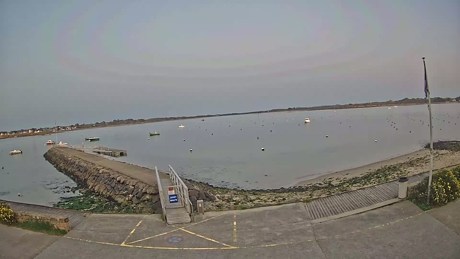 A calm, overcast day reveals a small harbor with several boats moored, a stone jetty extending into the water, and a ramp leading to a paved area.