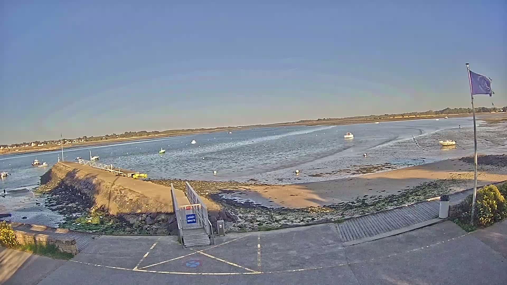 A small harbor with several boats, a stone pier with a wooden ramp, and a sandy shore under a clear blue sky.