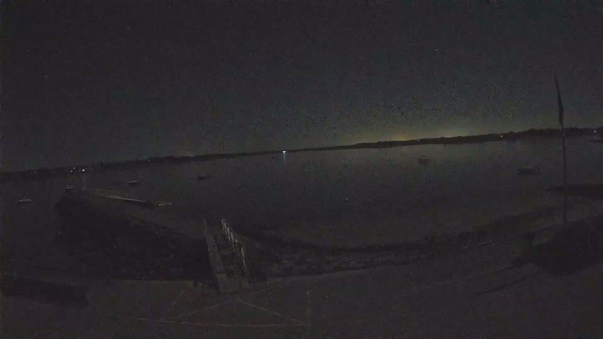 A nighttime view of a dark body of water with several boats, a shoreline, and a dark sky.