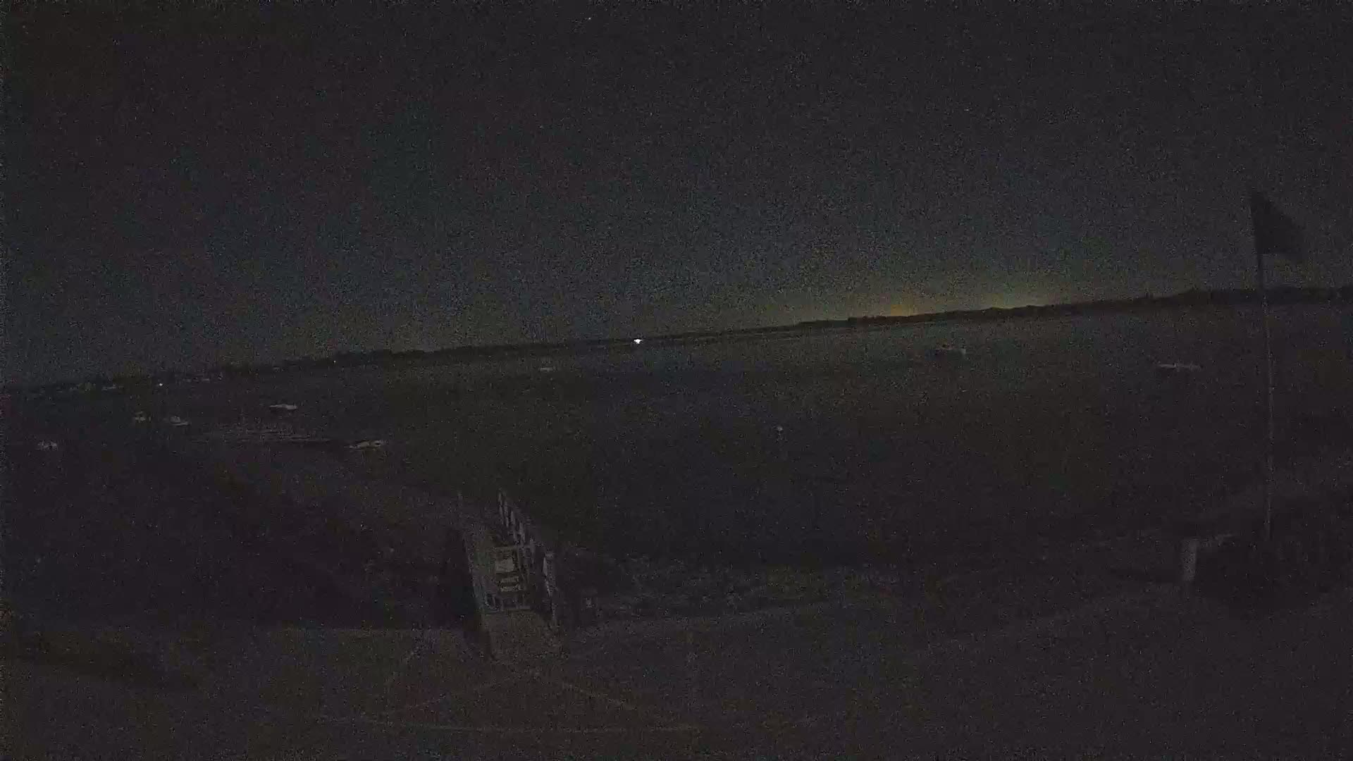A nighttime view of a dark body of water with a few indistinct objects, possibly boats, and a flagpole visible on the right, under a mostly clear, dark sky.