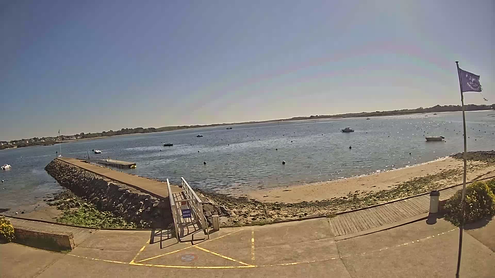 A stone jetty extends into a calm body of water under a clear blue sky, with several small boats scattered across the bay and a sandy shore nearby.