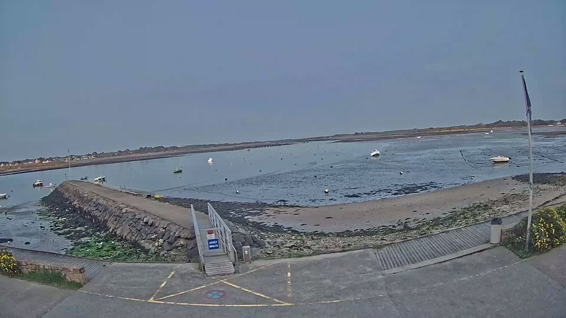 A calm, overcast day reveals a rocky, low-tide shoreline with several small boats scattered in the water and a wooden ramp leading to a stone jetty.