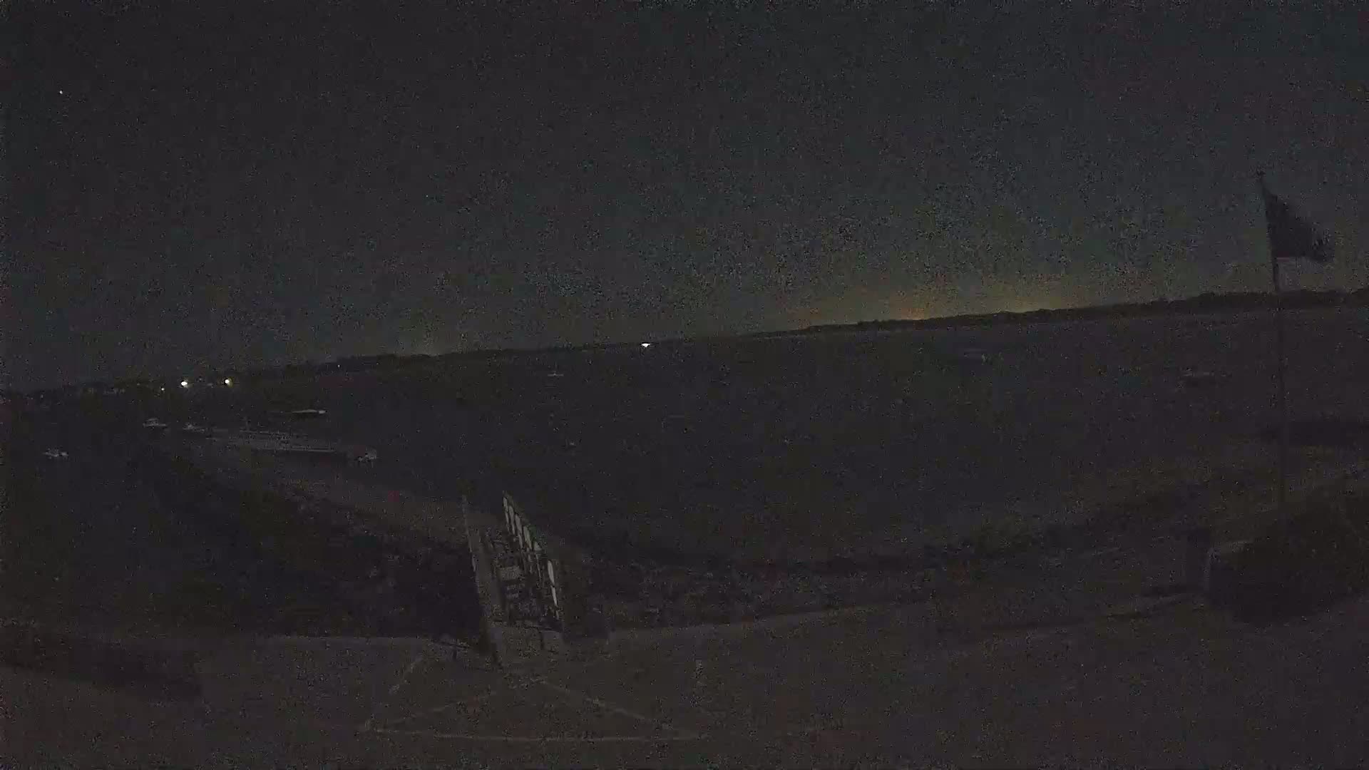 A nighttime, low-light view of a dark, flat expanse of land near water, with a flagpole visible in the upper right corner and faint lights in the distance.