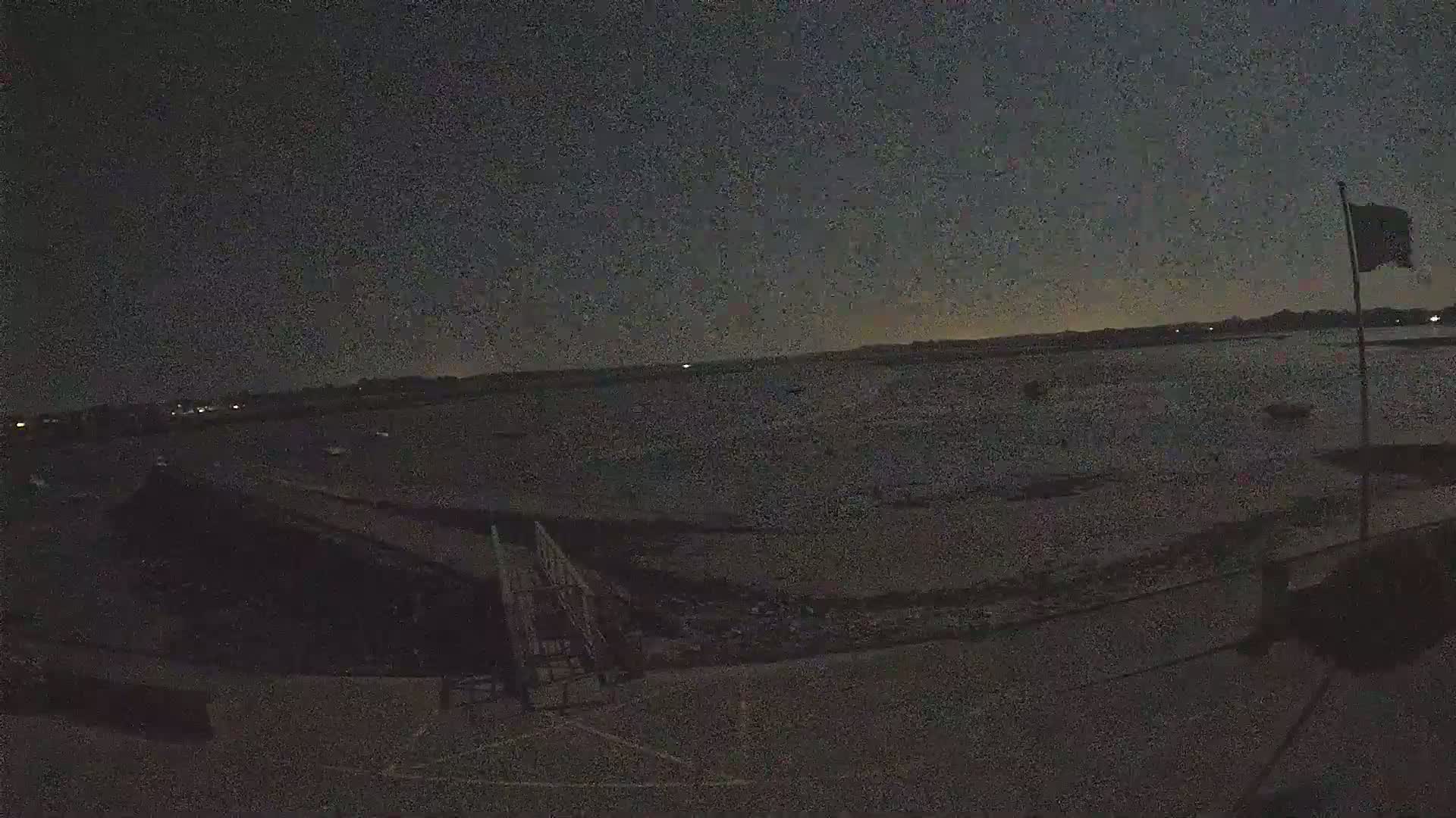 A dark, nighttime view of a body of water with a few boats visible in the distance, a small structure and ramp in the foreground, and a flagpole with a flag on the right.