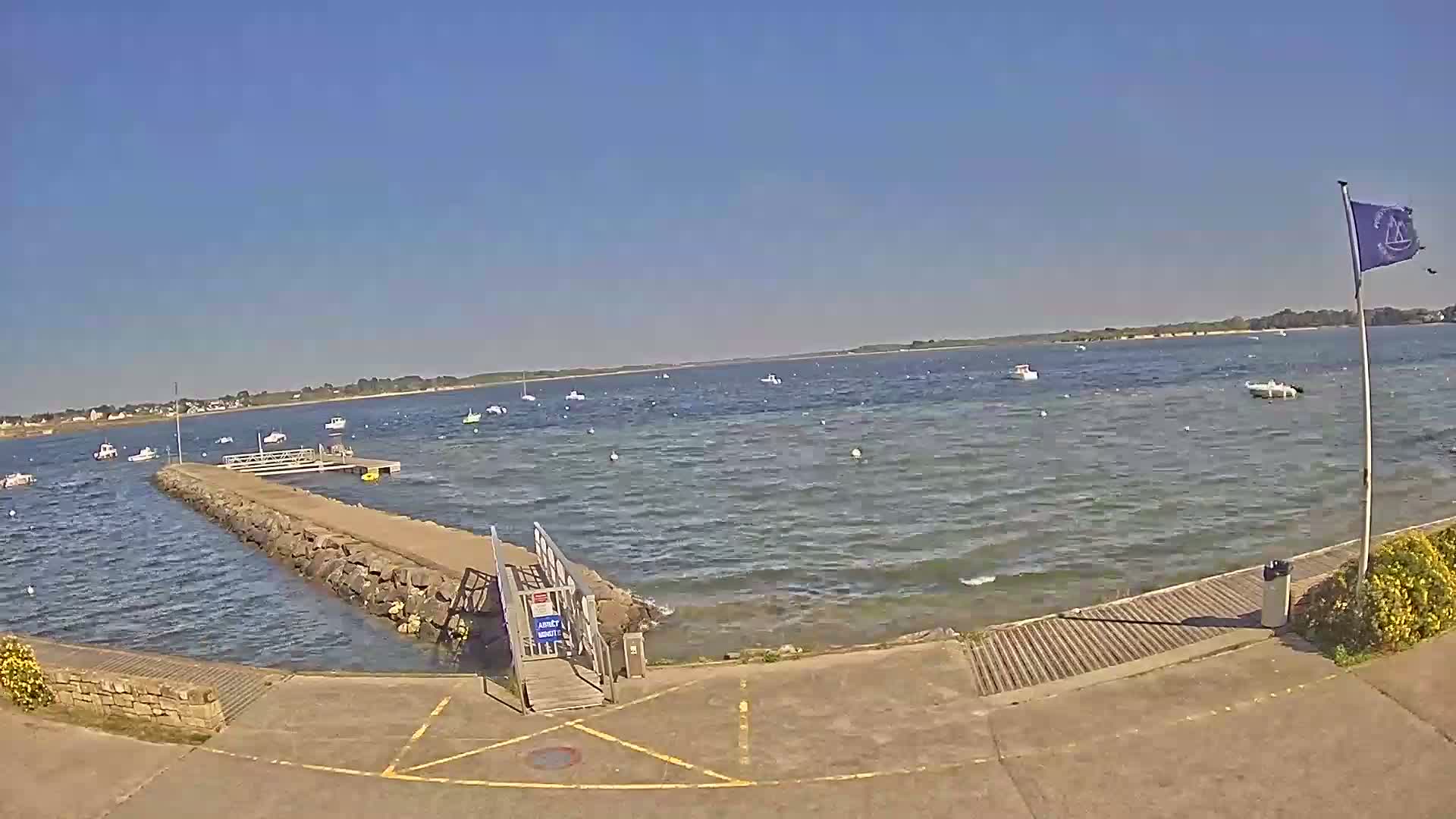 A stone jetty extends into a calm body of water dotted with small boats under a clear blue sky.