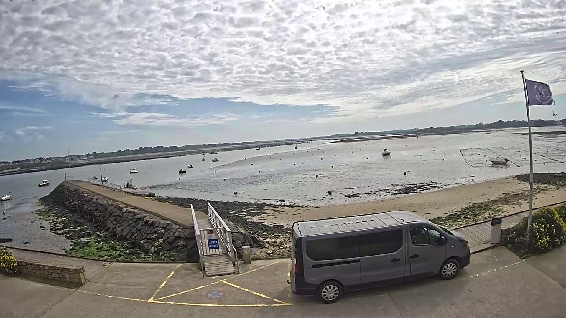 A gray minivan is parked near a stone pier overlooking a calm bay with numerous small boats and oyster beds under a partly cloudy sky.