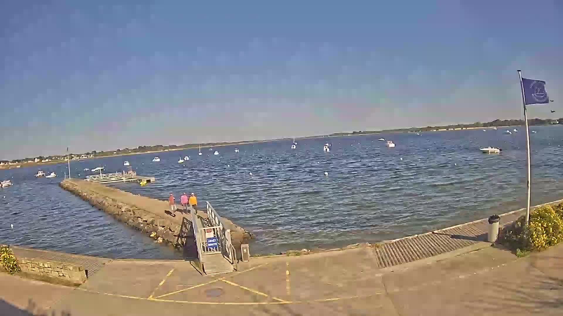 A calm, sunny day reveals a tranquil body of water dotted with numerous small boats, a stone pier with people walking on it, and a ramp leading to a paved area.