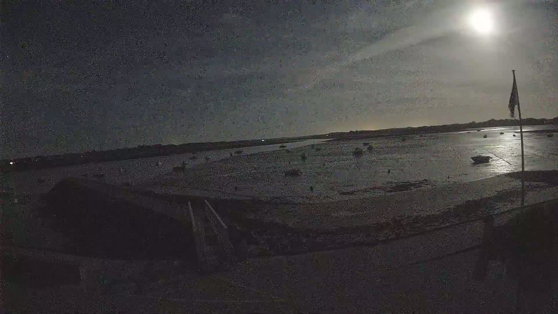 A bright moon illuminates a mostly flat, dark shoreline with several small boats visible in the shallow water at night.