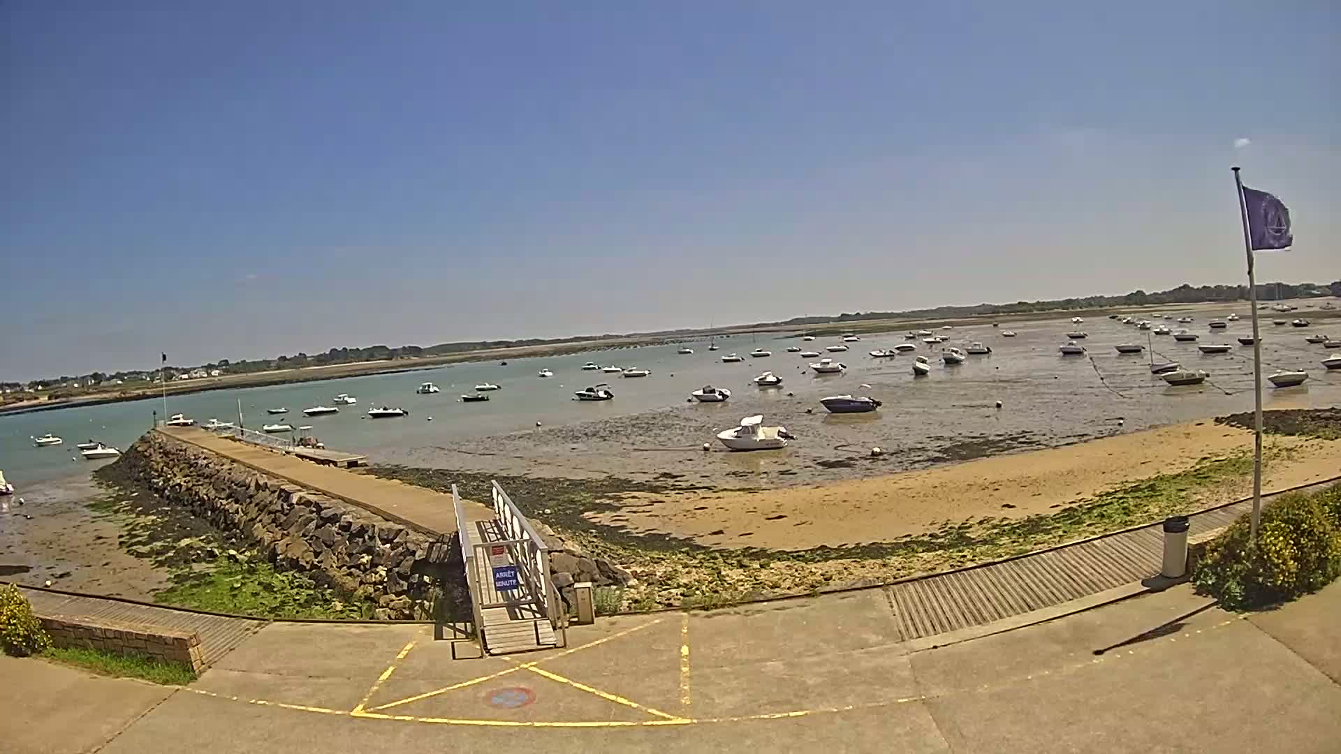 A sunny day reveals a calm body of water with numerous small boats moored at low tide near a stone jetty and sandy beach.