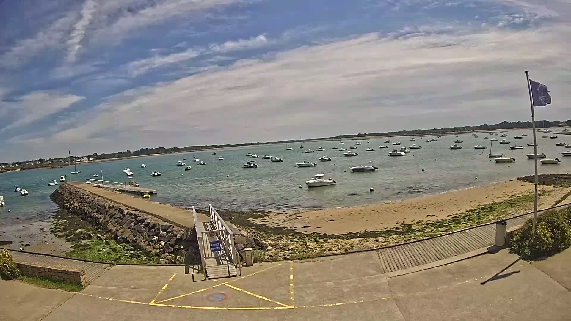 A sunny day with partly cloudy skies shows a calm bay with numerous small boats moored near a stone jetty and sandy shore.