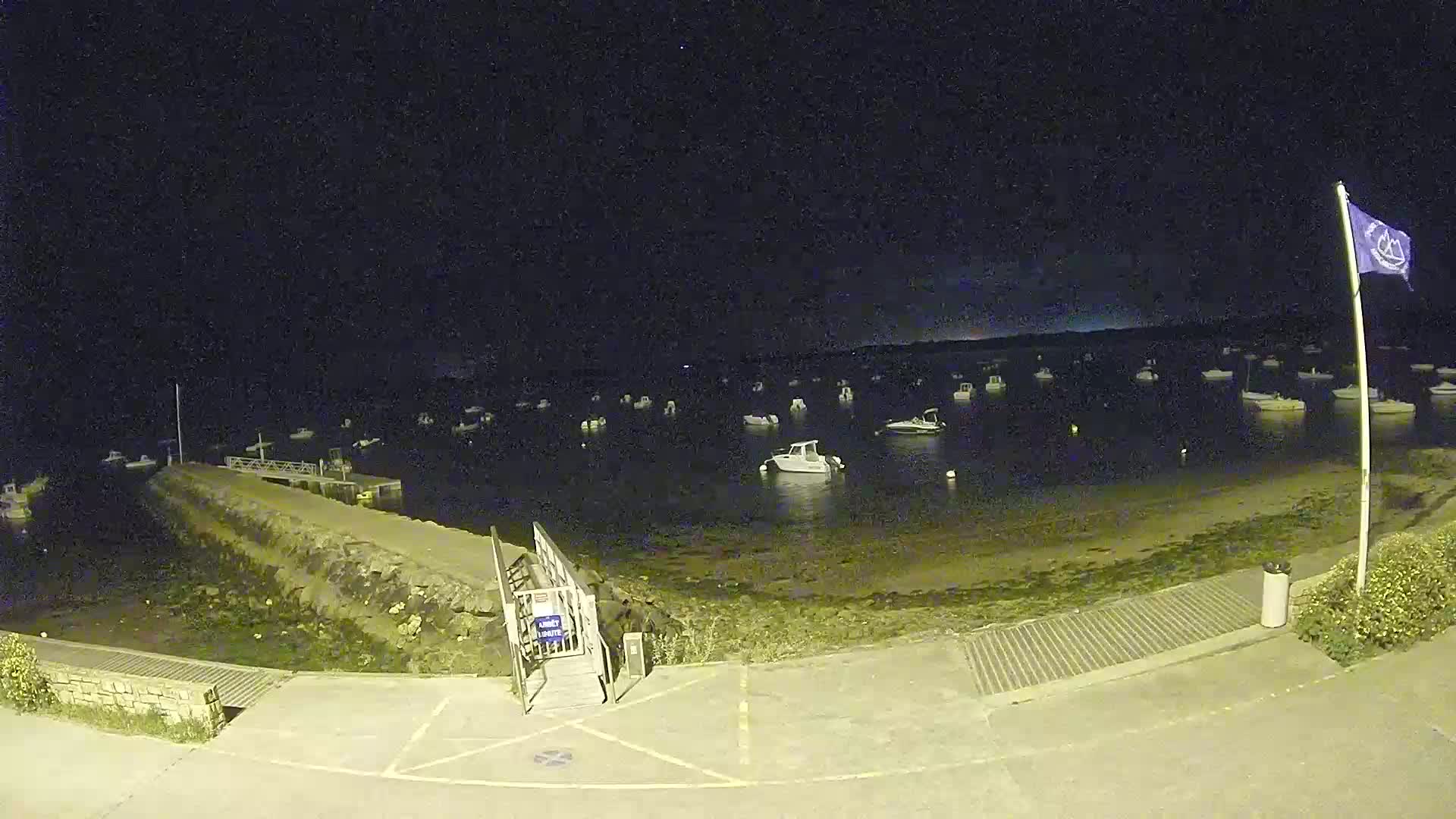 At night, a calm body of water with numerous boats is seen from a dock, under a dark sky.