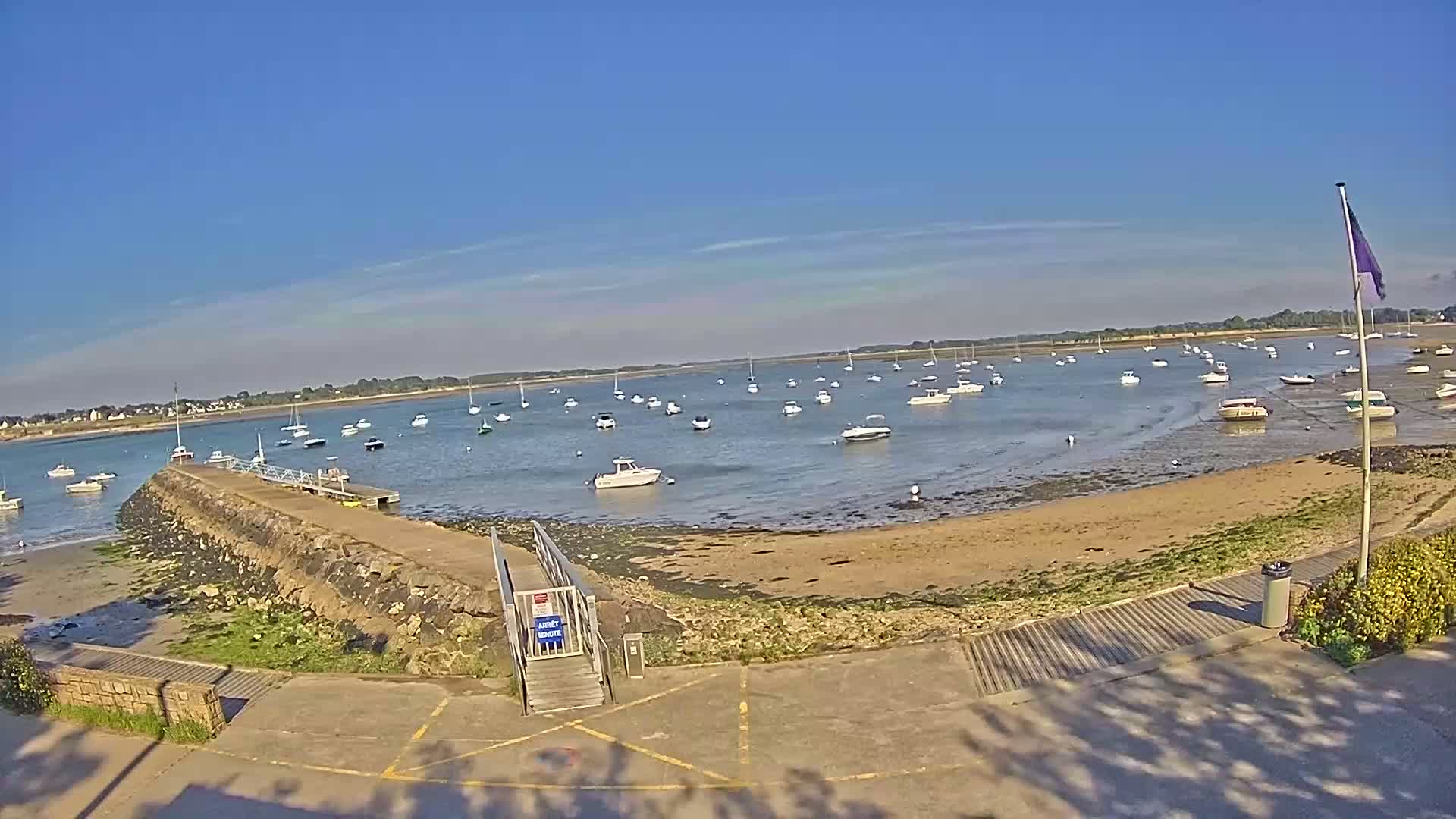 A calm, sunny day reveals a sandy shore with numerous boats at anchor in a bay, a stone jetty leading to a ramp, and a flagpole with a purple flag.