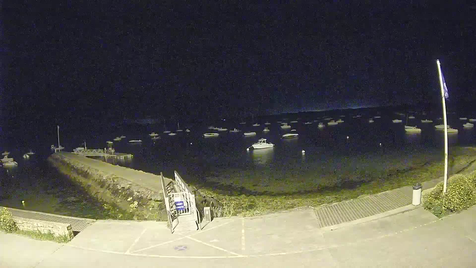 A nighttime view of a harbor with numerous boats at anchor, a small pier, and a paved area.