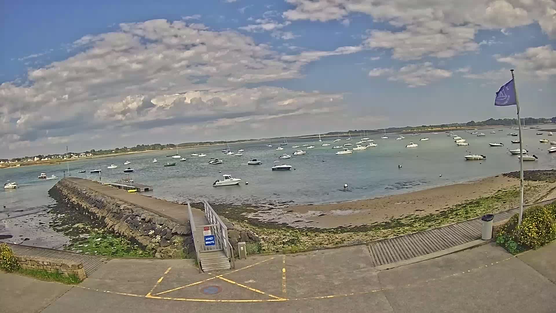 A partly cloudy day shows many small boats anchored in calm water near a stone jetty and sandy shore.