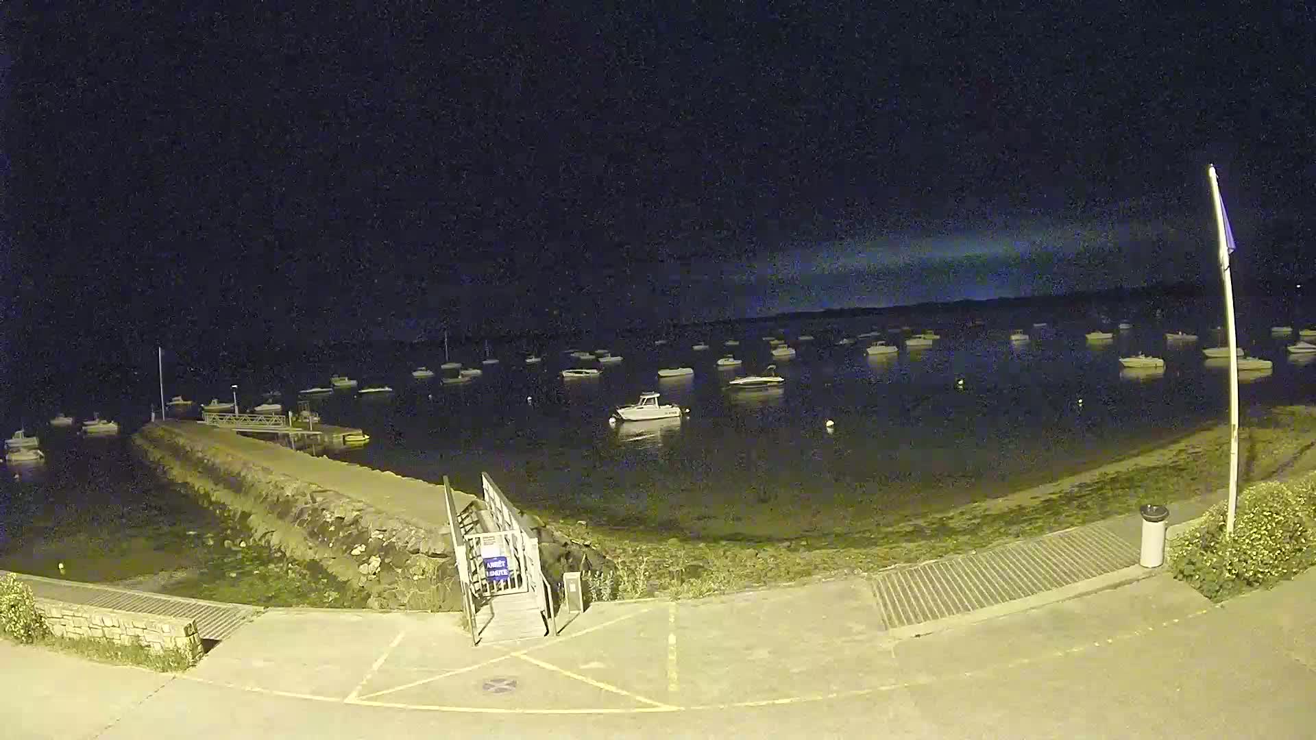 A nighttime view of a calm harbor with numerous boats moored, a small pier with a ramp, and a paved area.