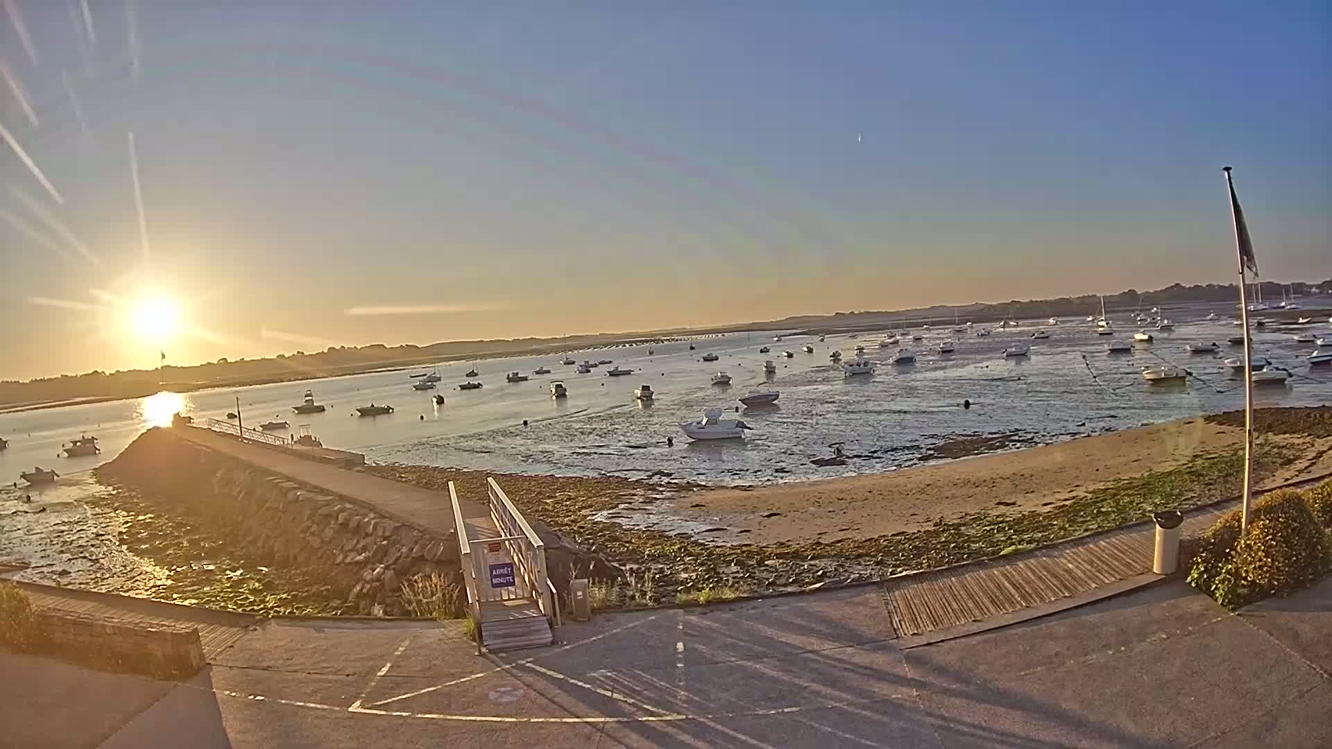 A calm, sun-drenched bay at sunset, filled with numerous small boats moored near a stone jetty and sandy shore.