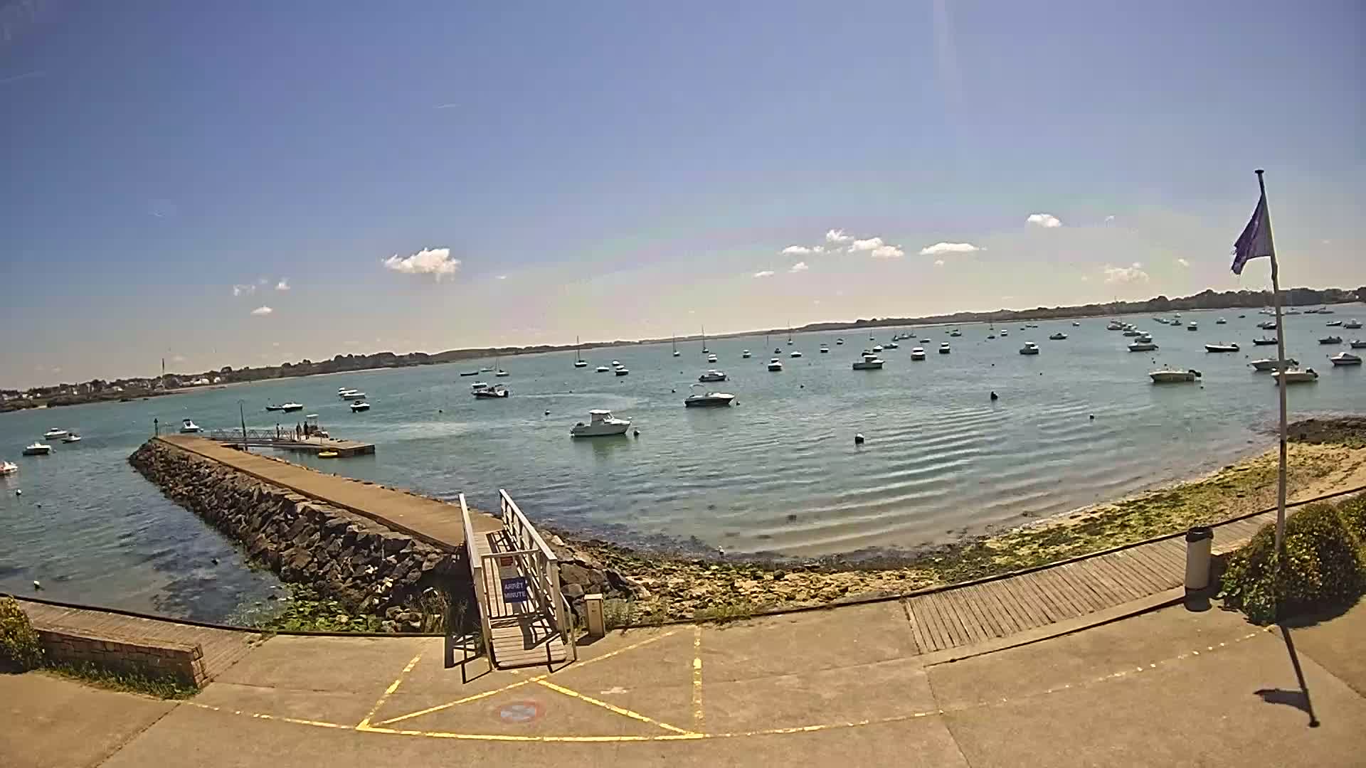 A sunny day shows a calm bay with numerous boats at anchor, a stone jetty leading to a small pier, and a paved area with a wooden walkway nearby.