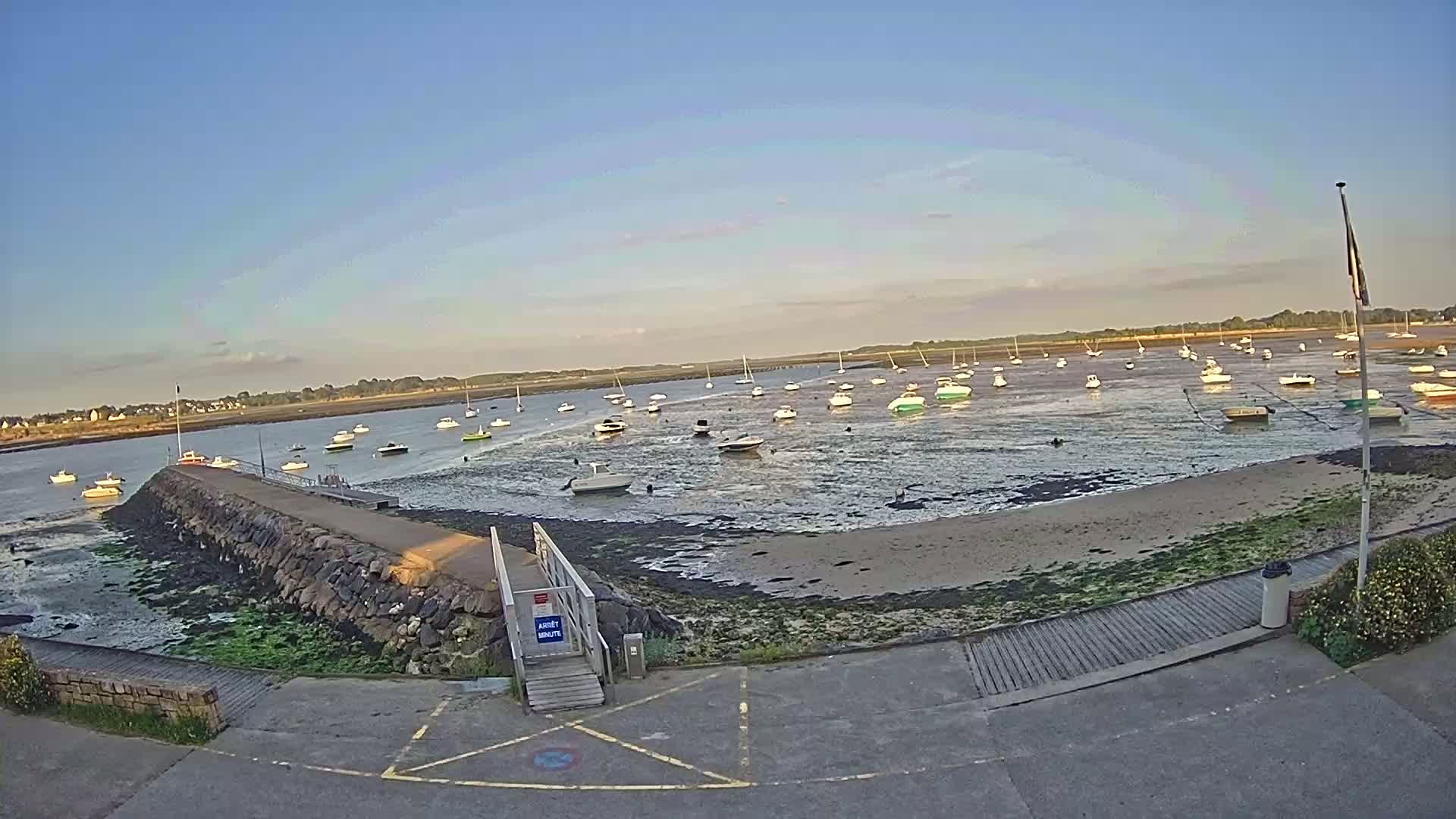 Many small boats are moored in shallow, calm water at low tide near a stone jetty on a sunny day.