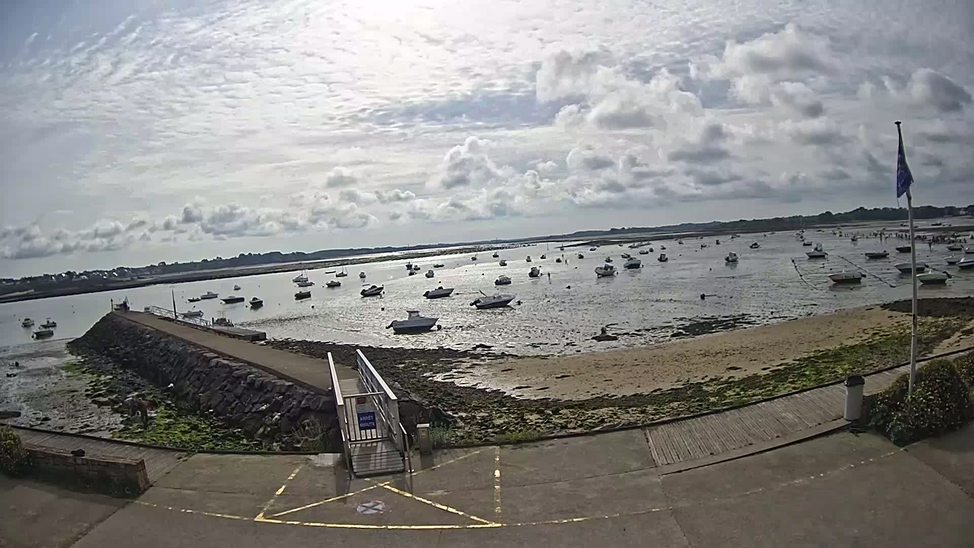 A partly cloudy day reveals a calm body of water with numerous small boats moored near a rocky shore and a paved walkway leading to a pier.