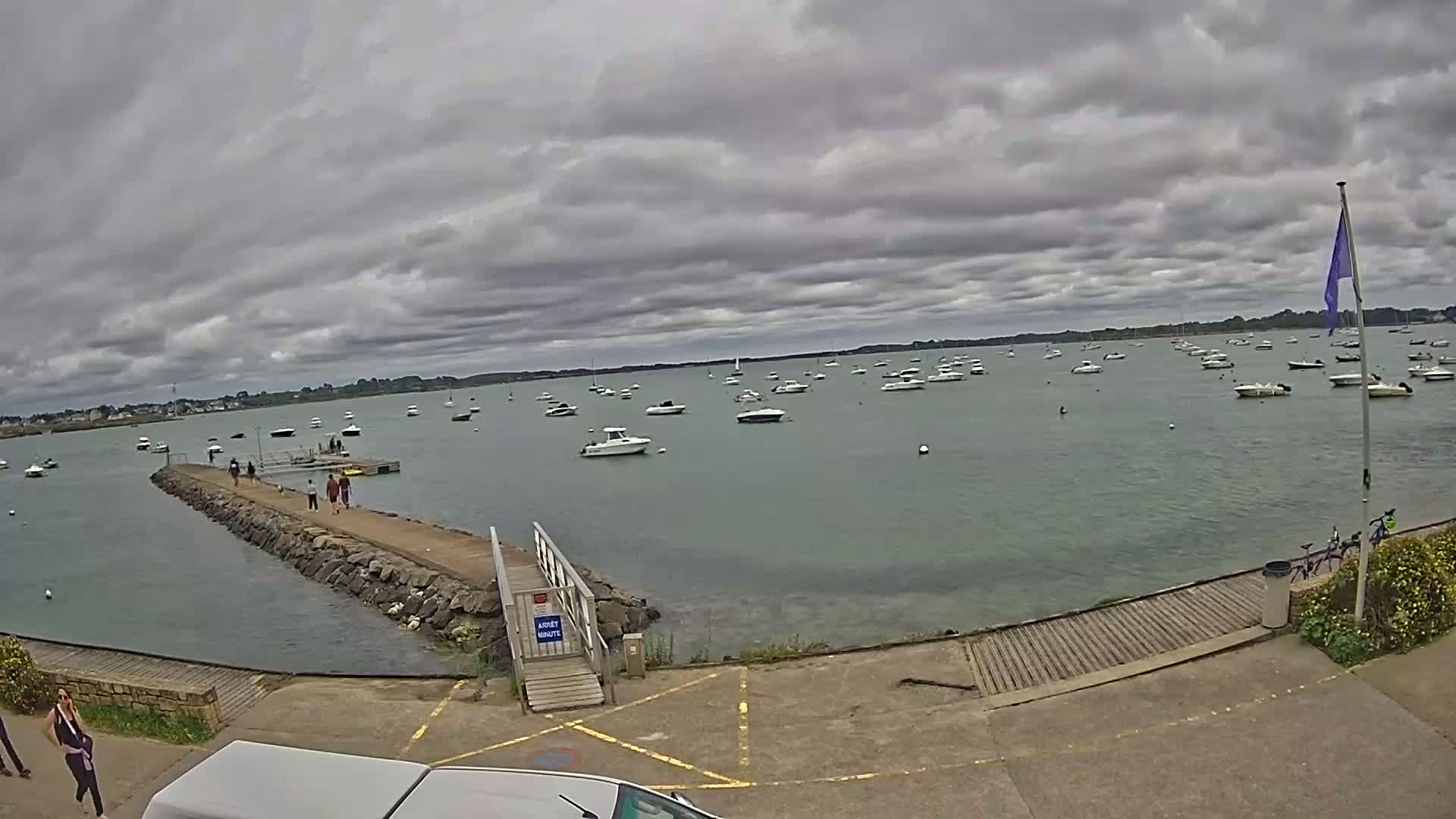 A calm body of water with numerous small boats is viewed from a rocky pier under an overcast sky.