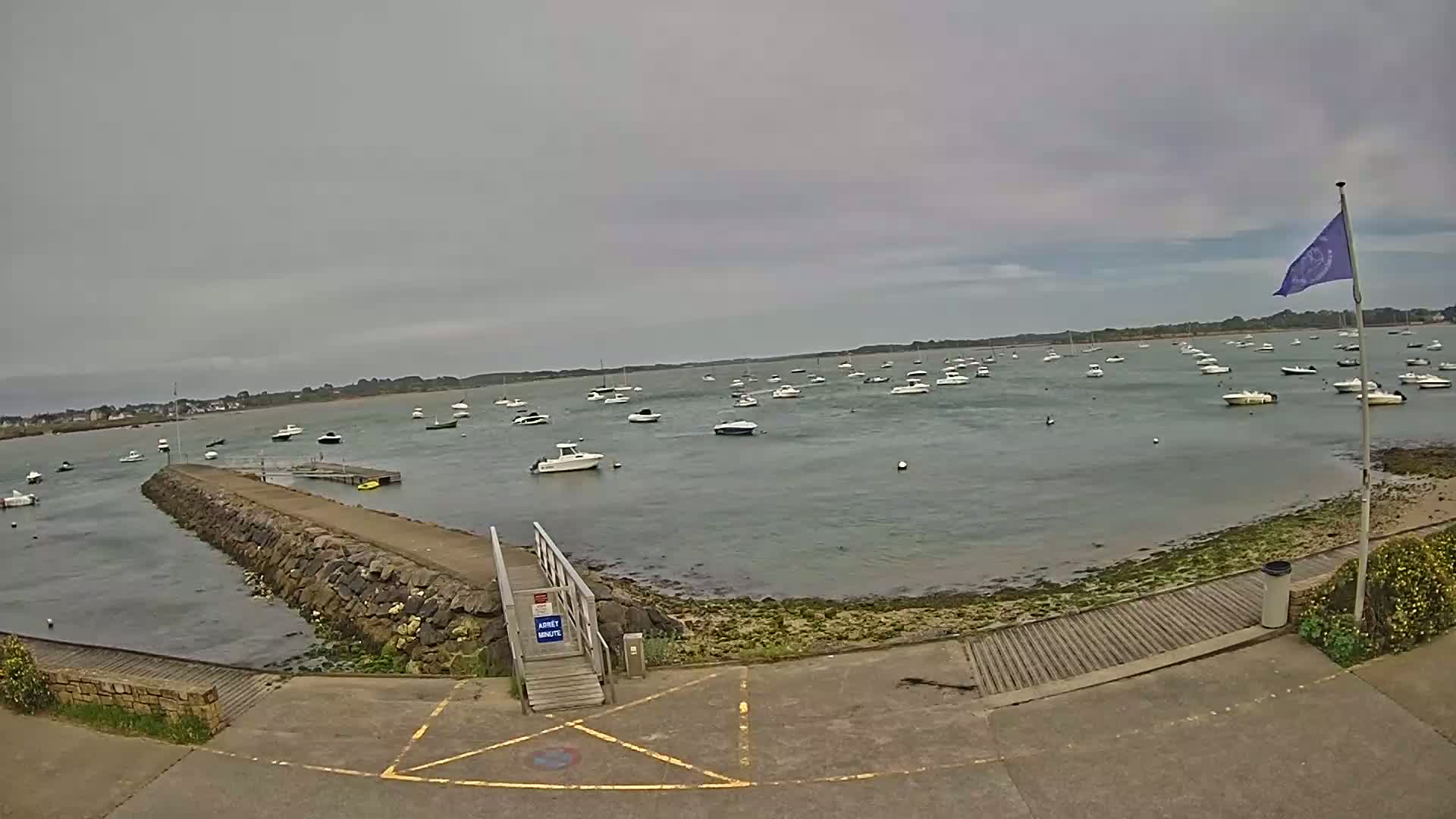 A calm, overcast day shows a harbor filled with numerous small motorboats moored near a stone jetty and a boat ramp.
