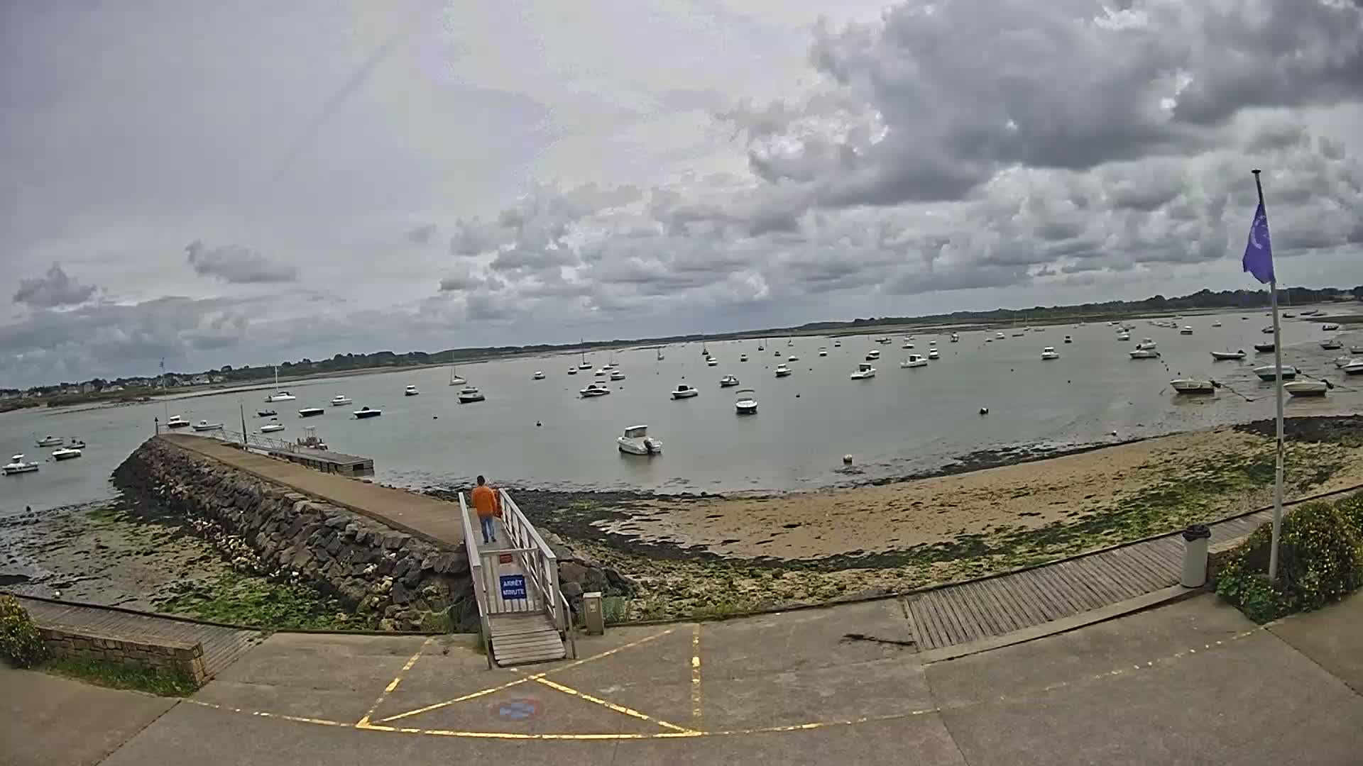 A person stands on a stone jetty overlooking a calm bay filled with numerous small boats under a cloudy sky.