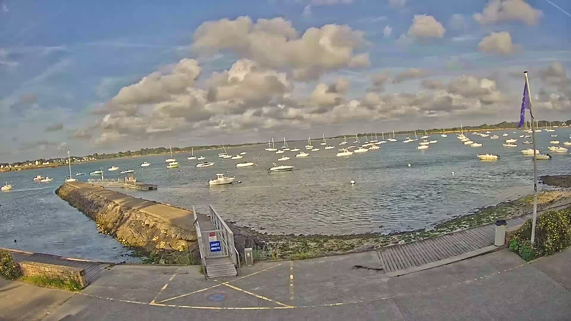 A mostly sunny day with puffy clouds over a calm body of water where numerous sailboats and motorboats are anchored near a stone jetty and ramp.