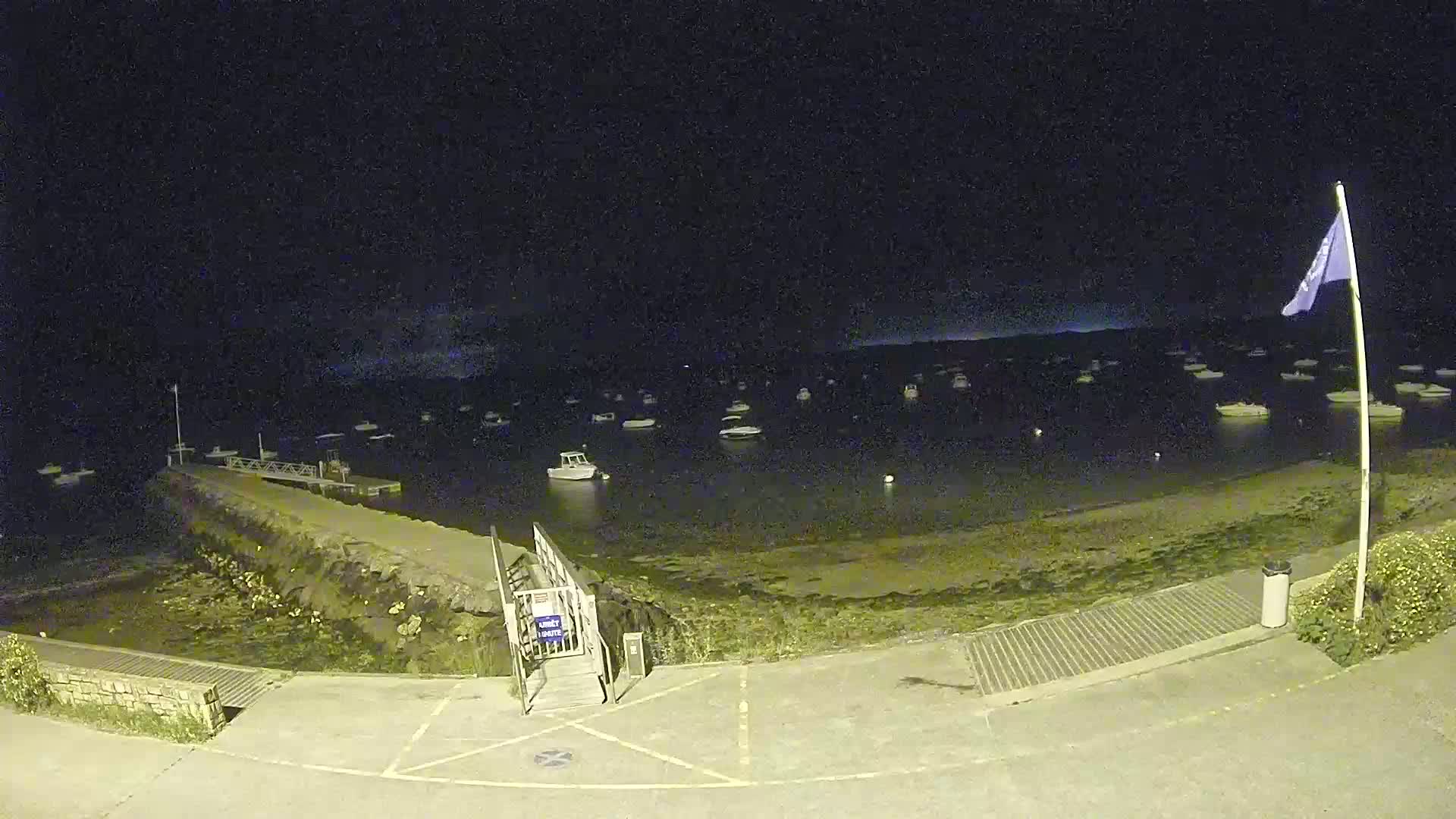 A nighttime view of a harbor with numerous boats at anchor, a small pier with a ramp, and a flagpole with a flag flying.