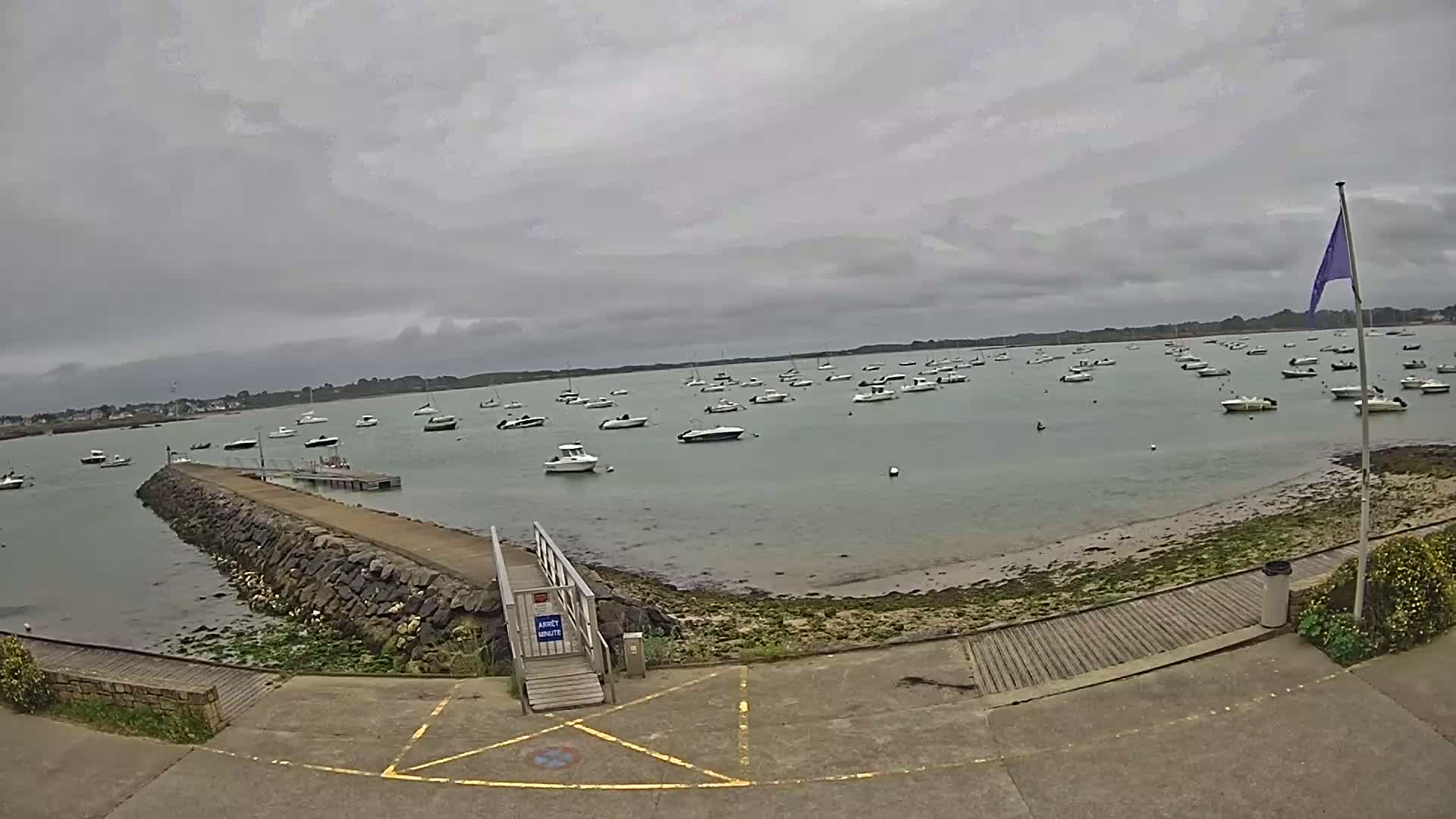 Numerous boats are moored in calm, gray-skied waters near a stone jetty and small wooden ramp.