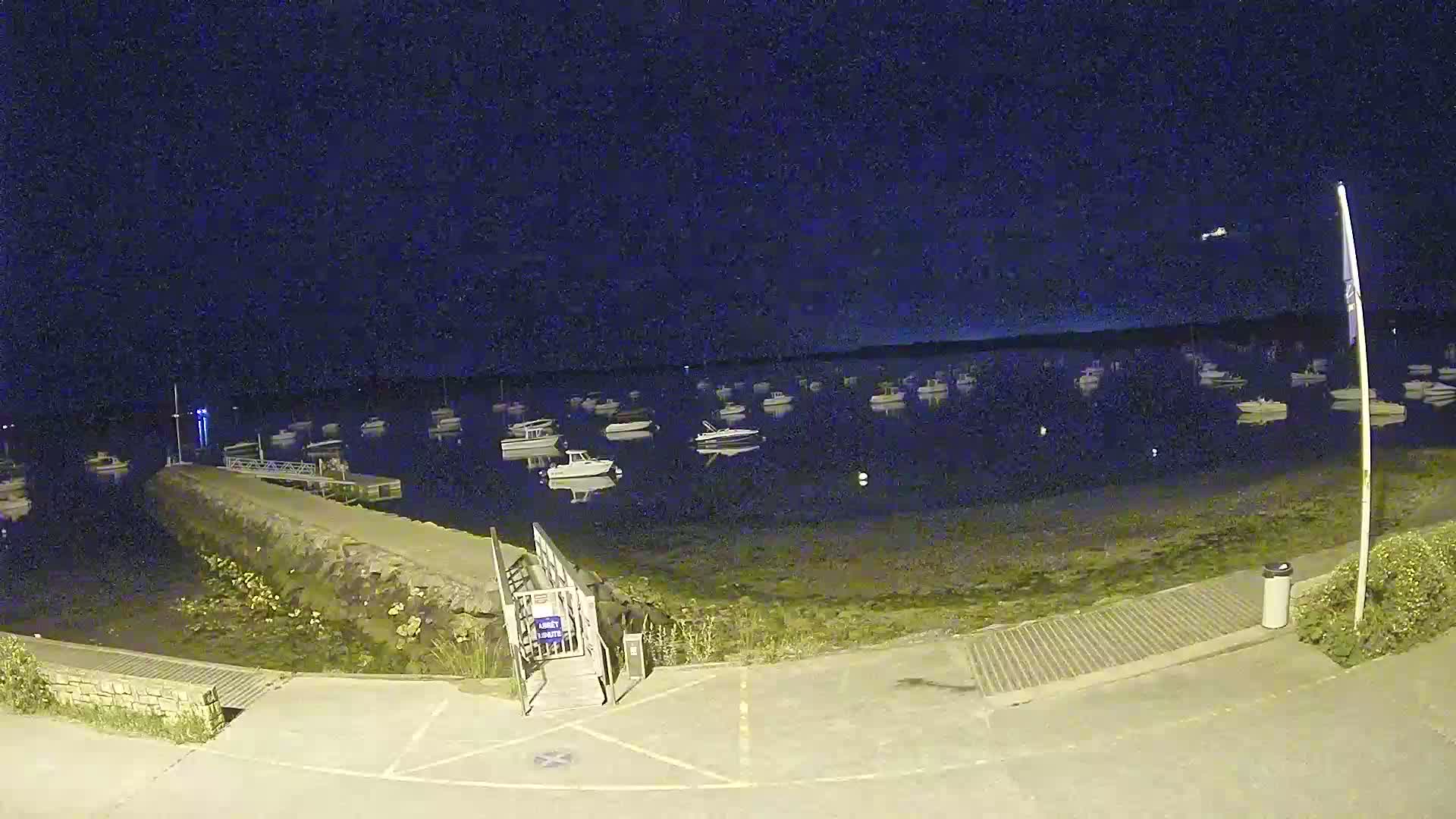 At night, a small harbor shows numerous boats moored in calm water, with a ramp and paved area leading to a stone wall at the water's edge.