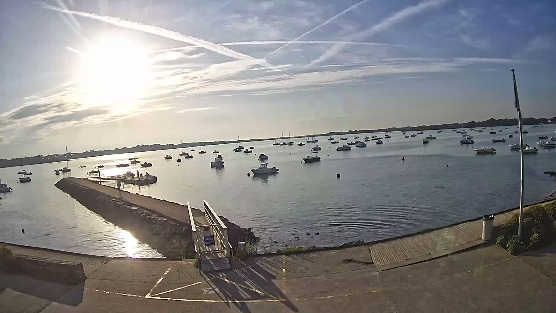 A calm body of water with numerous boats is viewed from a paved area leading to a pier under a mostly sunny sky with contrails.