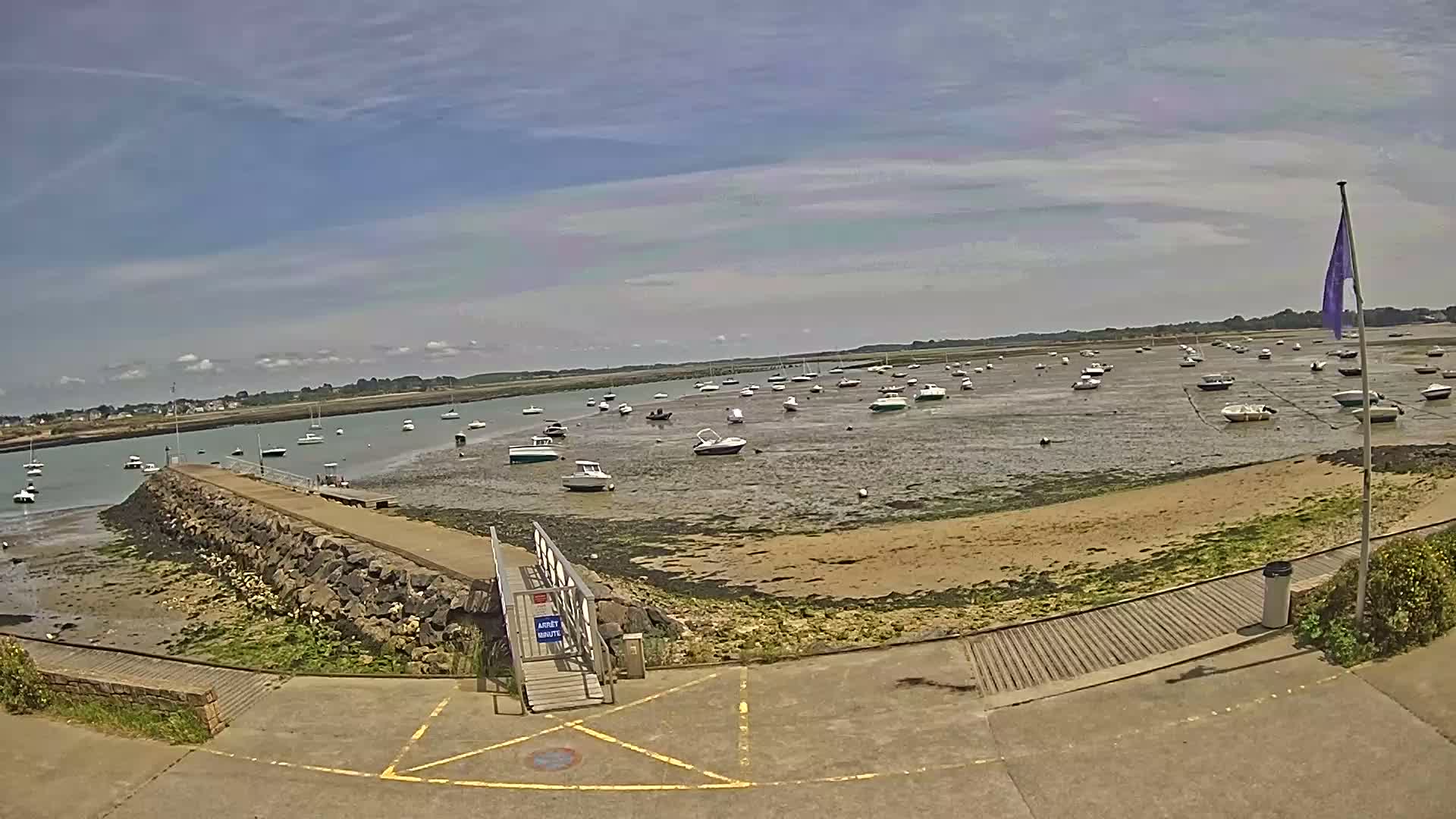 A mostly sunny day reveals numerous boats resting on a tidal flat near a stone jetty and paved walkway.
